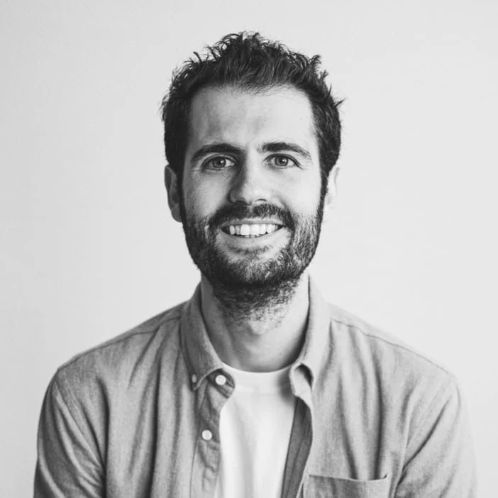 Portrait en noir et blanc d'un homme souriant portant une chemise et un t-shirt, avec une barbe et des cheveux courts en arrière-plan blanc.