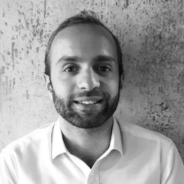 Portrait d'un homme avec une barbe, portant une chemise blanche, souriant devant un mur texturé.