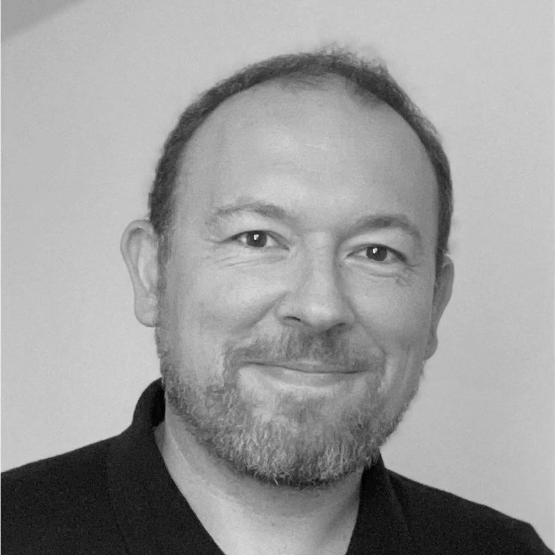 Un portrait en noir et blanc d'un homme avec une barbe et un sourire, portant une chemise noire.