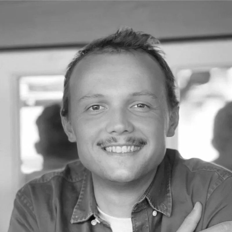 Portrait d'un homme souriant avec une moustache, portant une chemise en denim, dans un environnement intérieur lumineux.