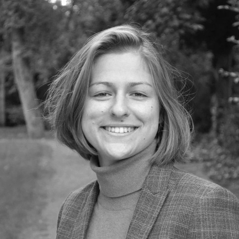 Photo en noir et blanc d'une jeune femme souriante avec des cheveux courts, portant un pull à col roulé et un blazer, en plein air avec des arbres en arrière-plan.
