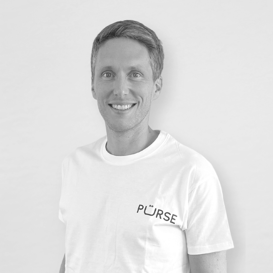 Jeune homme souriant portant un t-shirt blanc avec le mot 'PÜRSE' écrit dessus, debout contre un mur blanc.