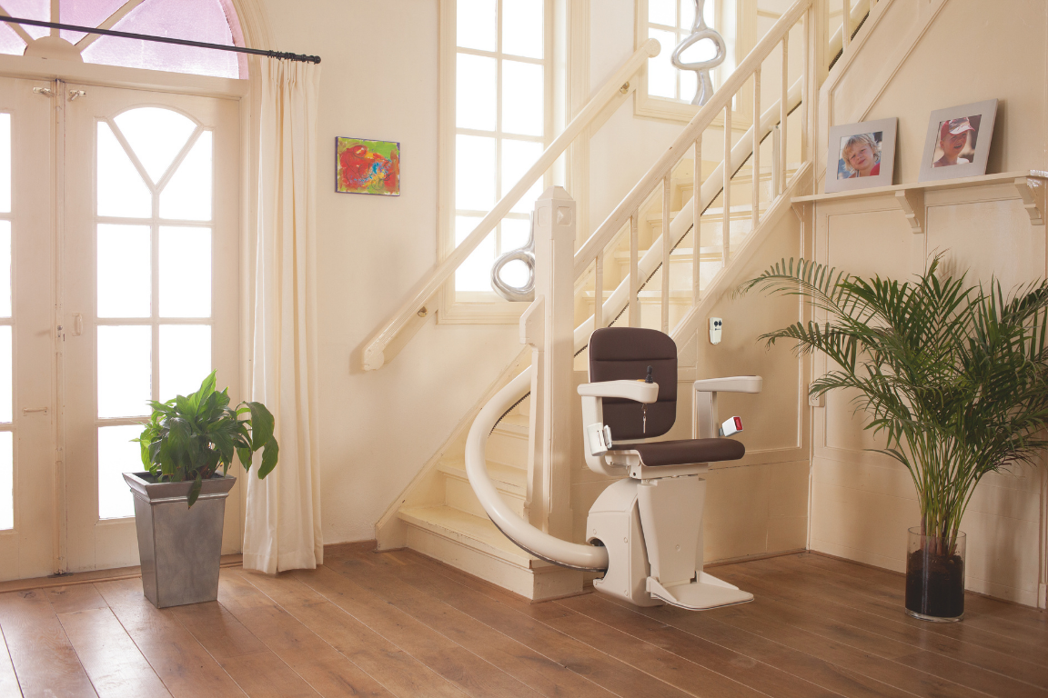 A staircase with a stairlift in a well-lit home interior, decorated with potted plants, framed photos, and a small colorful painting.