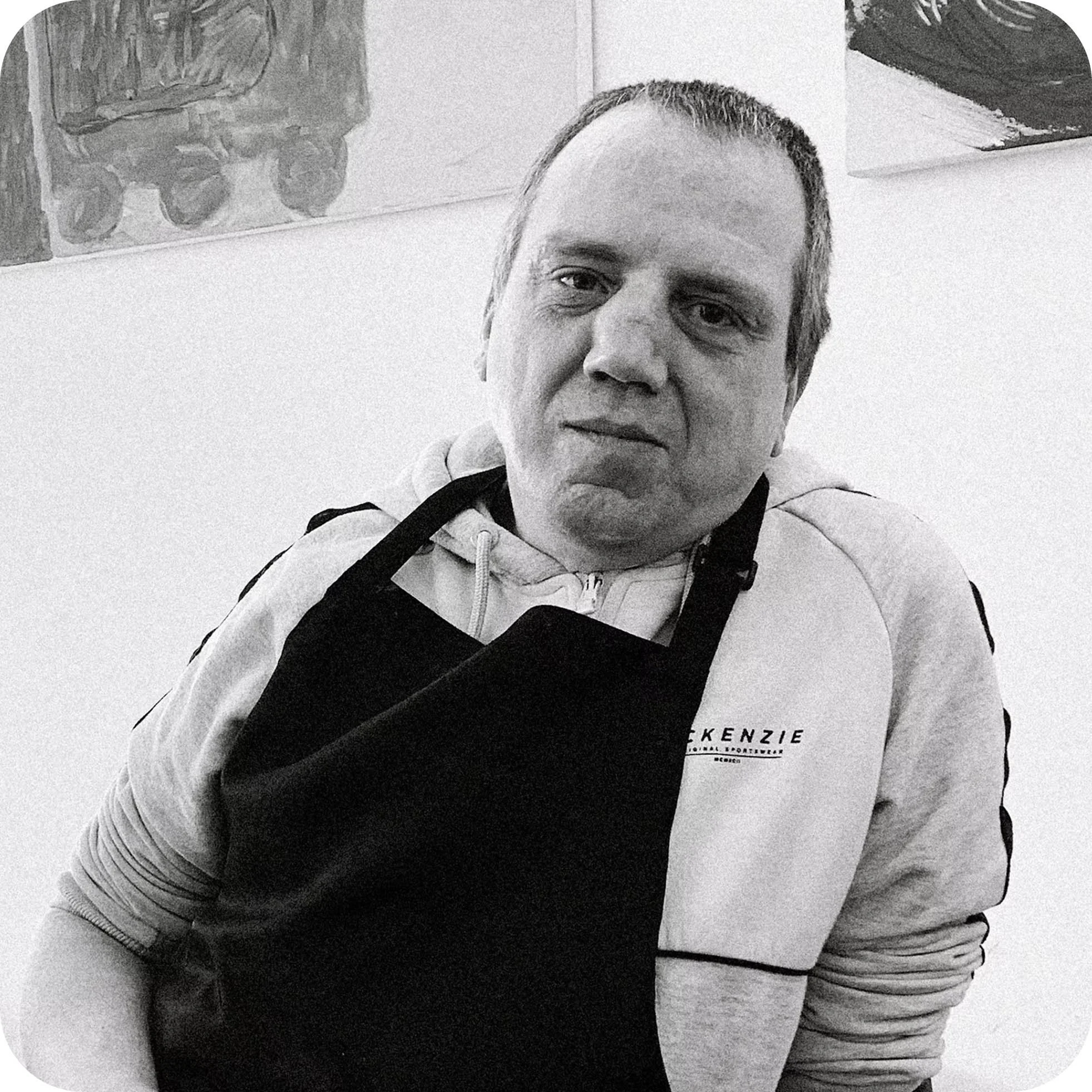 Black and white photo of a man with short hair, wearing a hoodie and a jacket, standing indoors near a white wall with abstract paintings hanging on it.