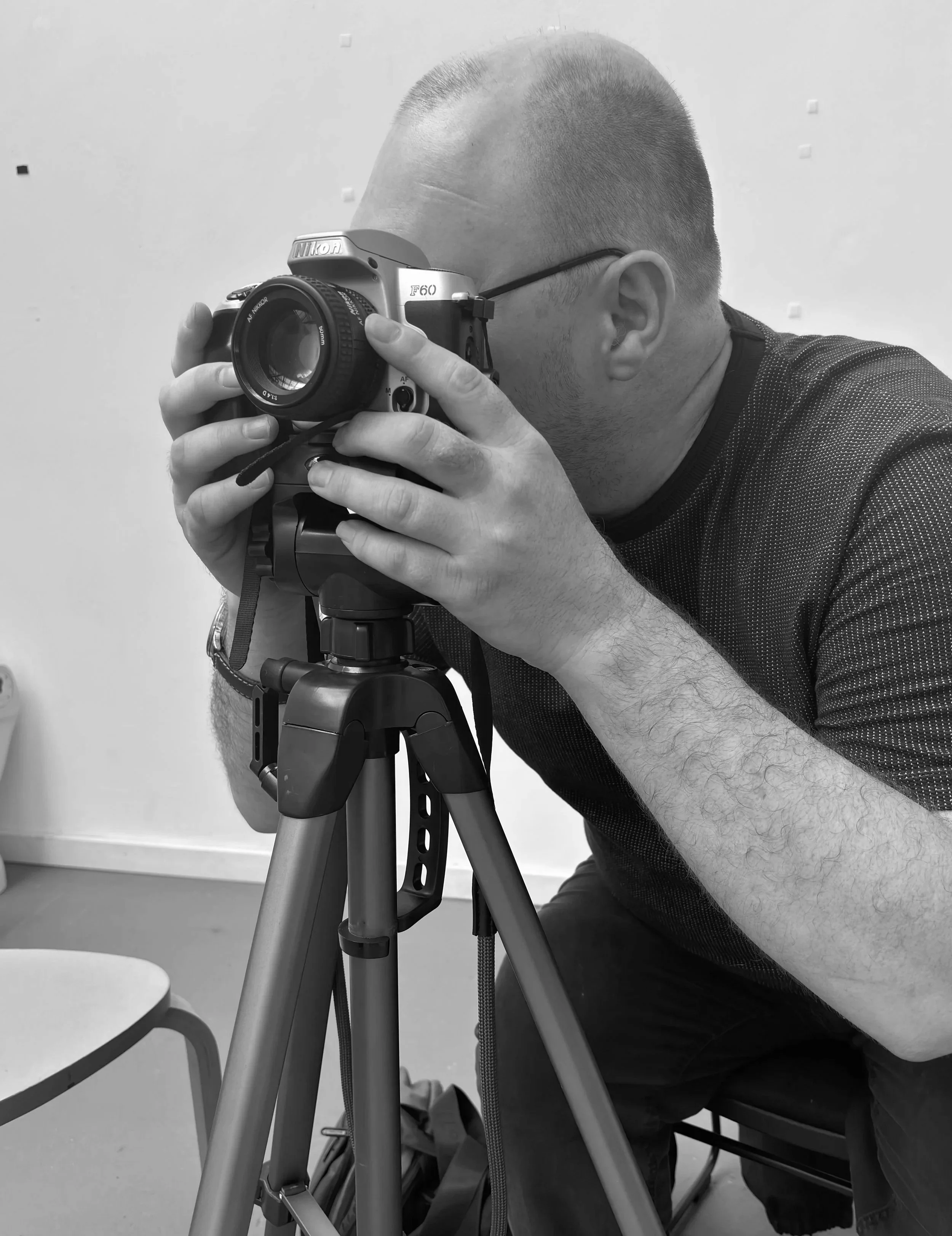 A man with a short haircut and glasses taking a photo with a Nikon camera mounted on a tripod, indoors.