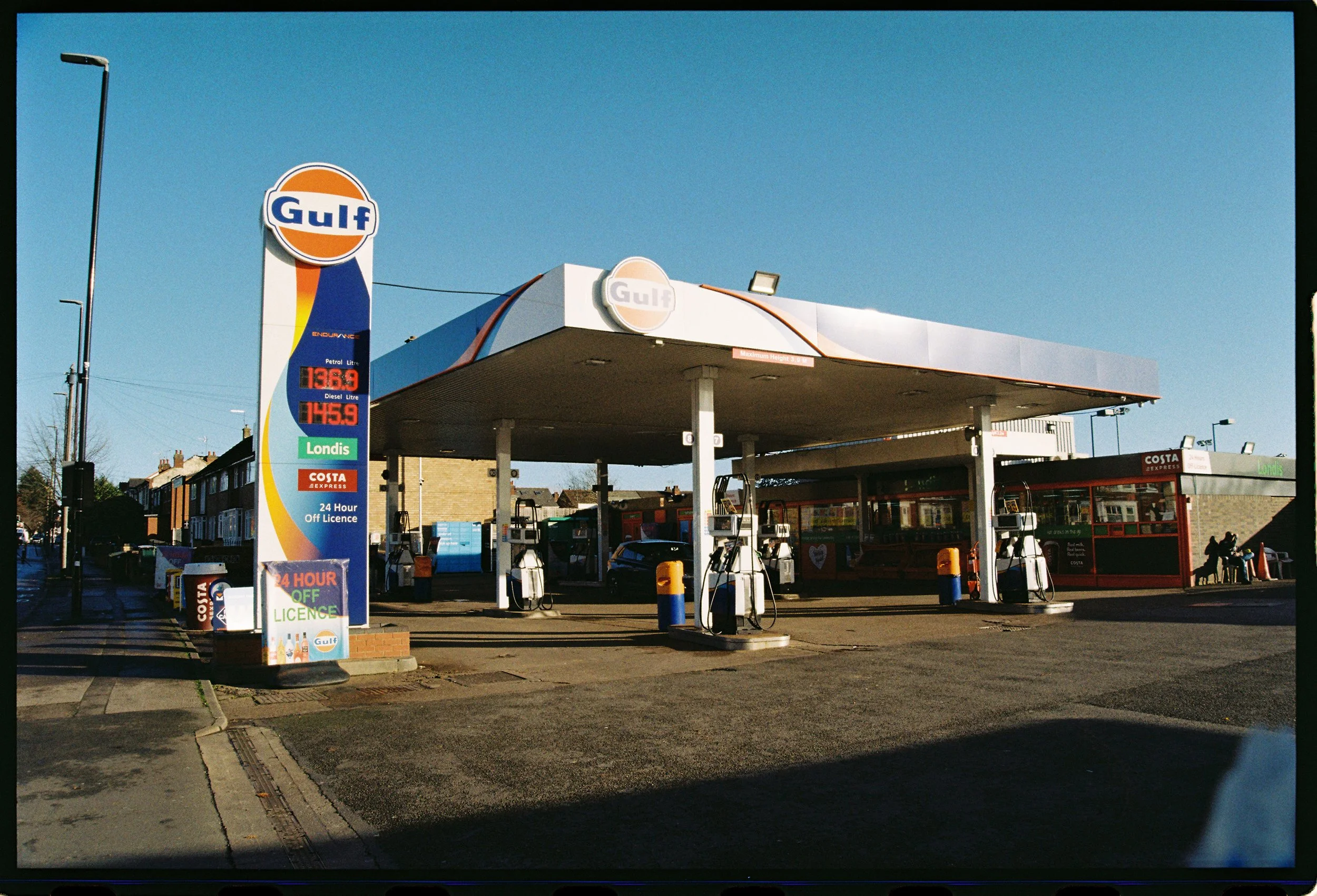 Gulf gas station with fuel prices displayed on a sign, featuring a blue sky, gas pumps under a canopy, and some buildings and trees in the background.