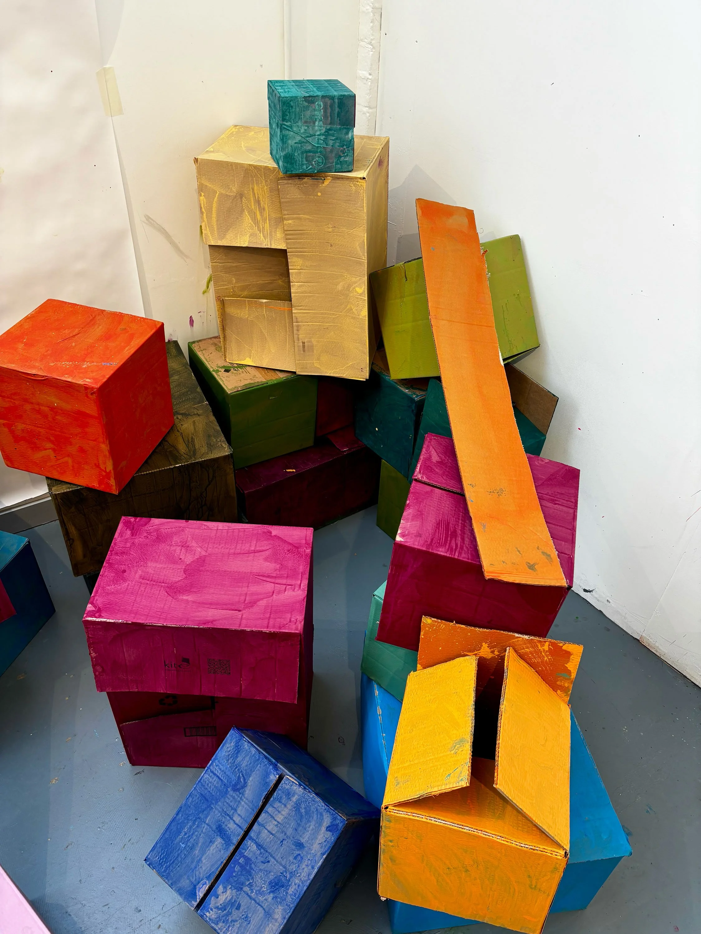 A pile of colorful cardboard boxes stacked unevenly against a white wall, with some boxes slightly tilted or leaning.
