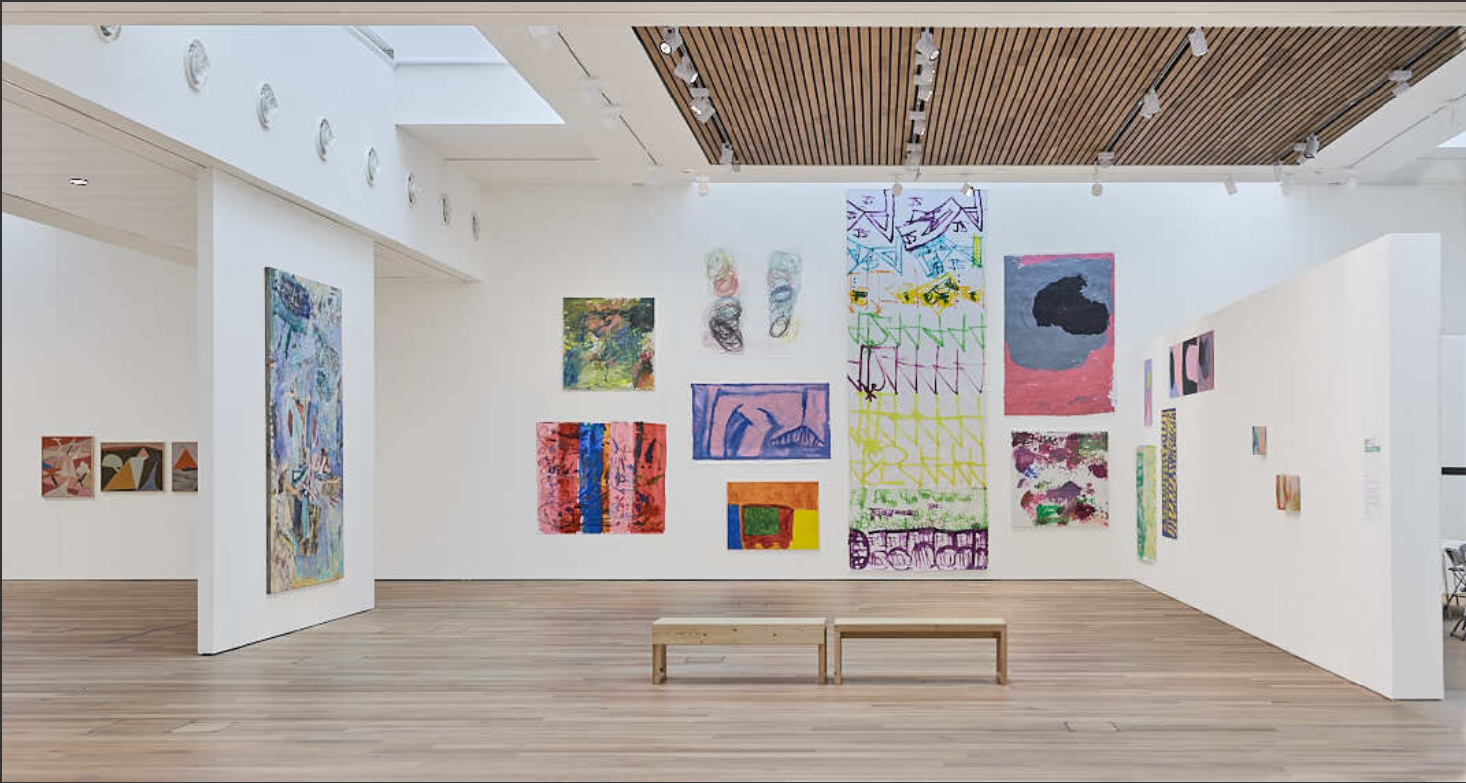 An art gallery with white walls displaying colorful abstract paintings and drawings, wooden flooring, and a wooden ceiling panel with track lighting. Two wooden benches are in the center of the room.