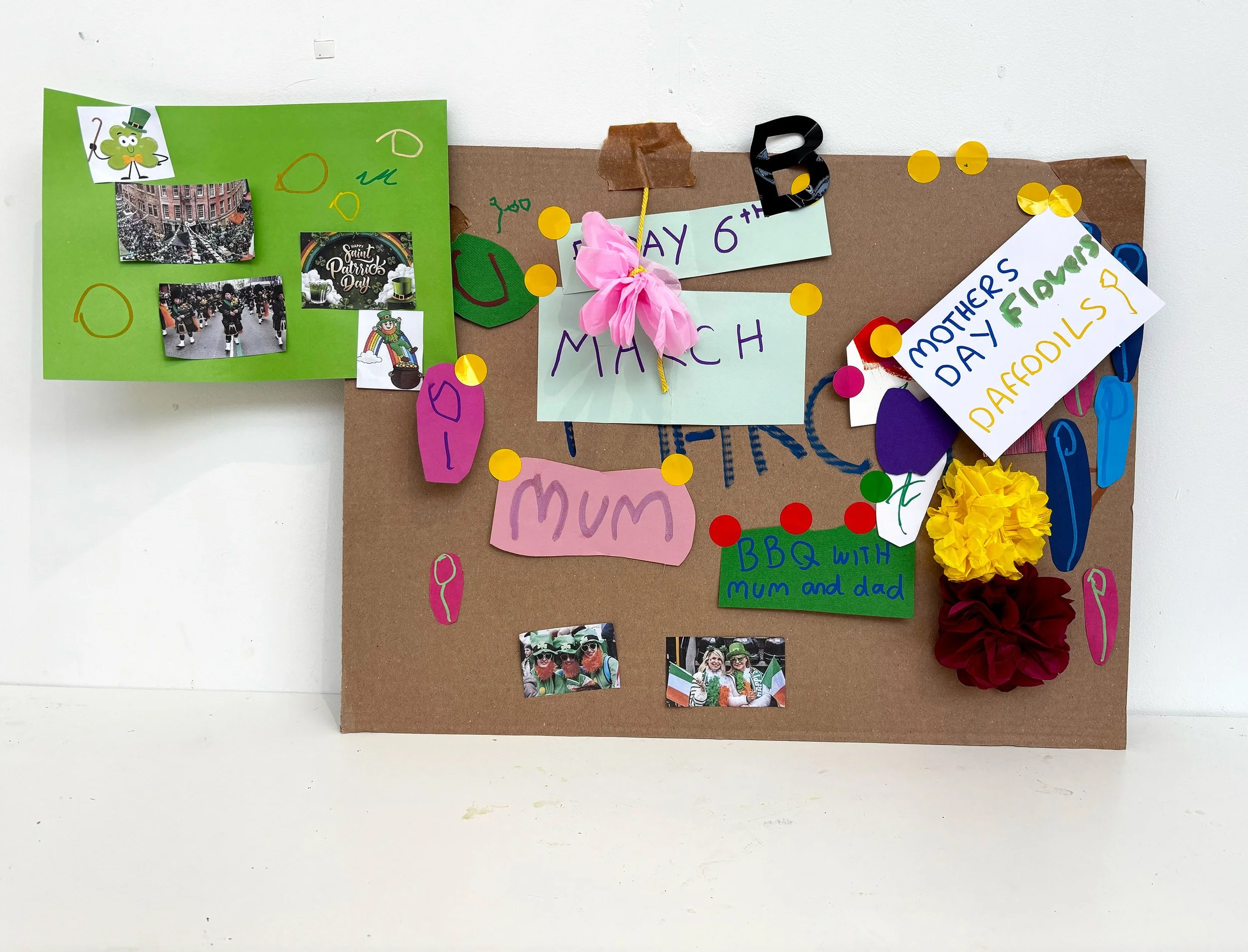 A decorated bulletin board for St. Patrick's Day featuring green and brown paper with photographs, drawings, and messages including "Happy St. Patrick's Day," "Mothers Day Flowers," and "BBQ with Mum and Dad." It also has shamrock and rainbow decorat
