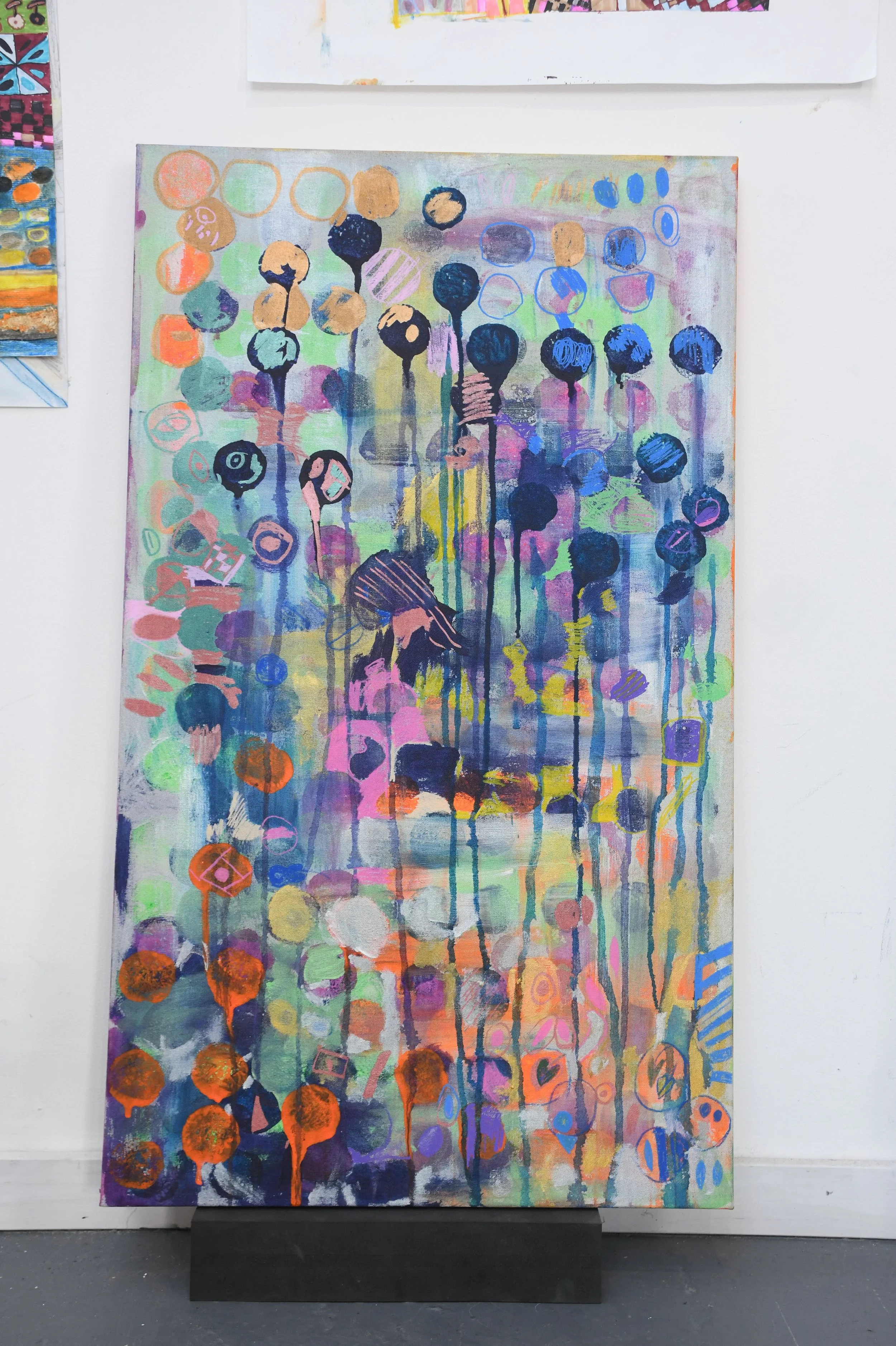 Abstract painting with multicolored circles, lines, and shapes on a canvas mounted on a black stand, displayed against a white wall.