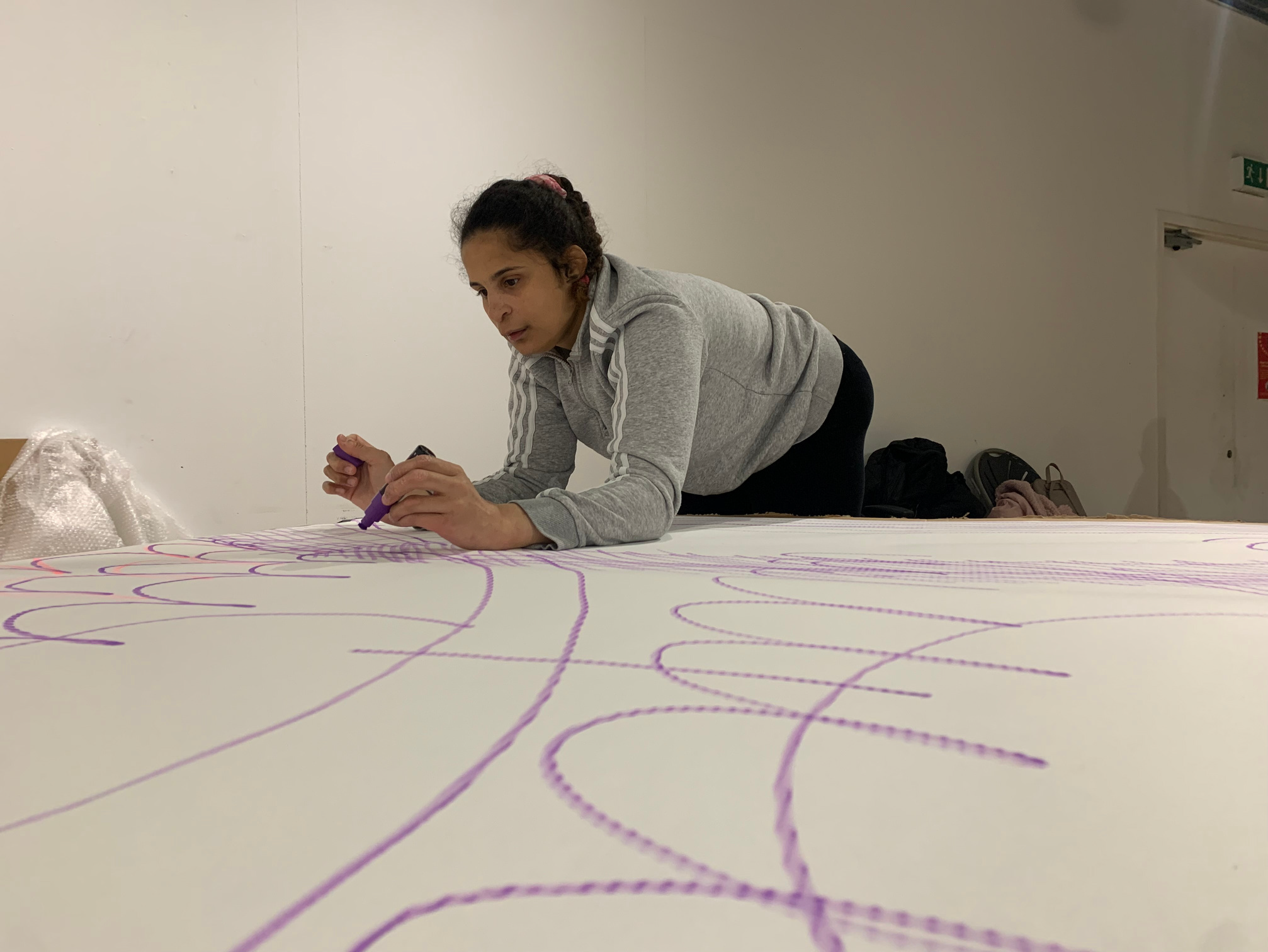 A young woman wearing a gray athletic hoodie and black leggings drawing with purple marker on large white paper spread on a table, in an indoor setting.