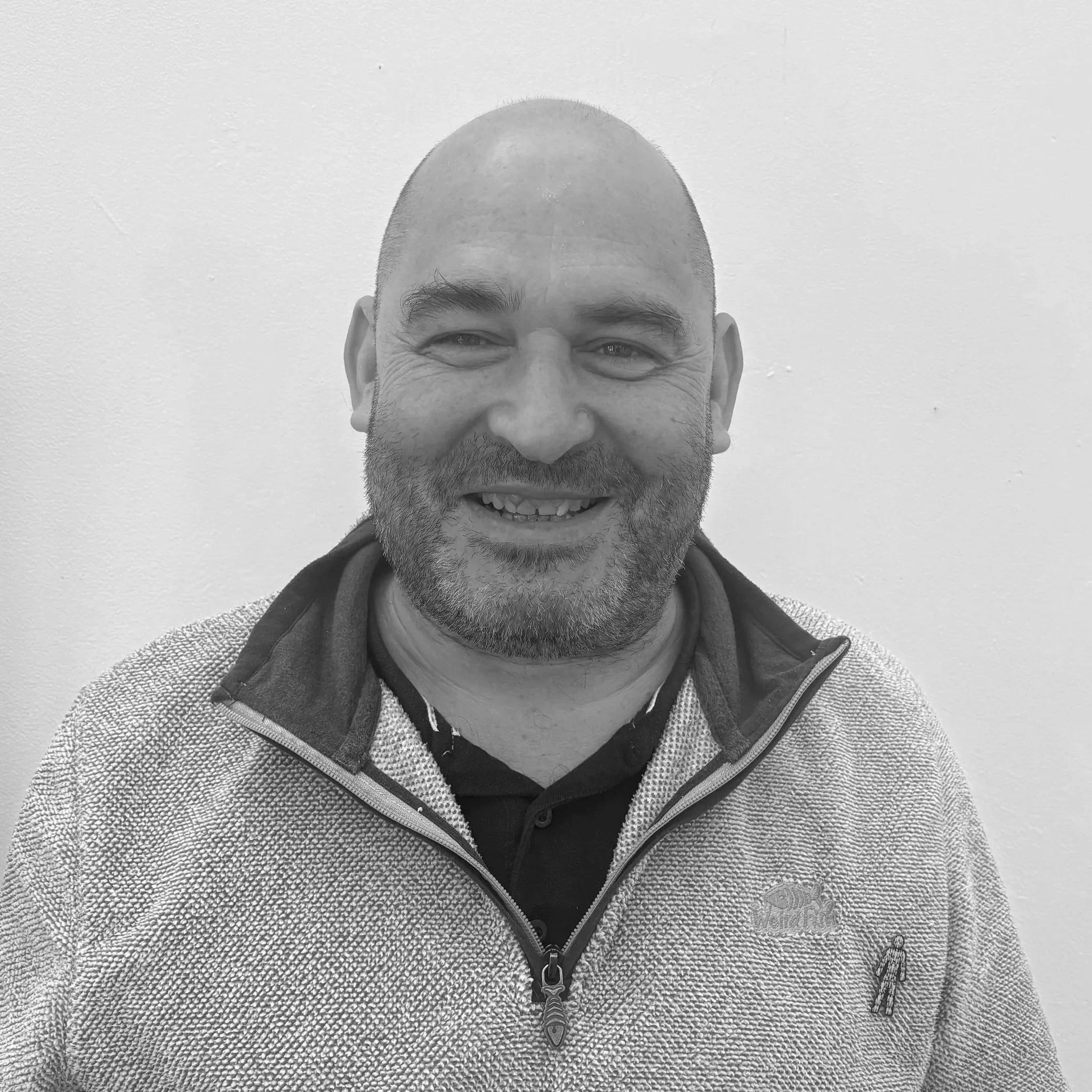 Man with a shaved head and beard smiling, wearing a patterned fleece jacket and a hoodie, standing against a plain wall.