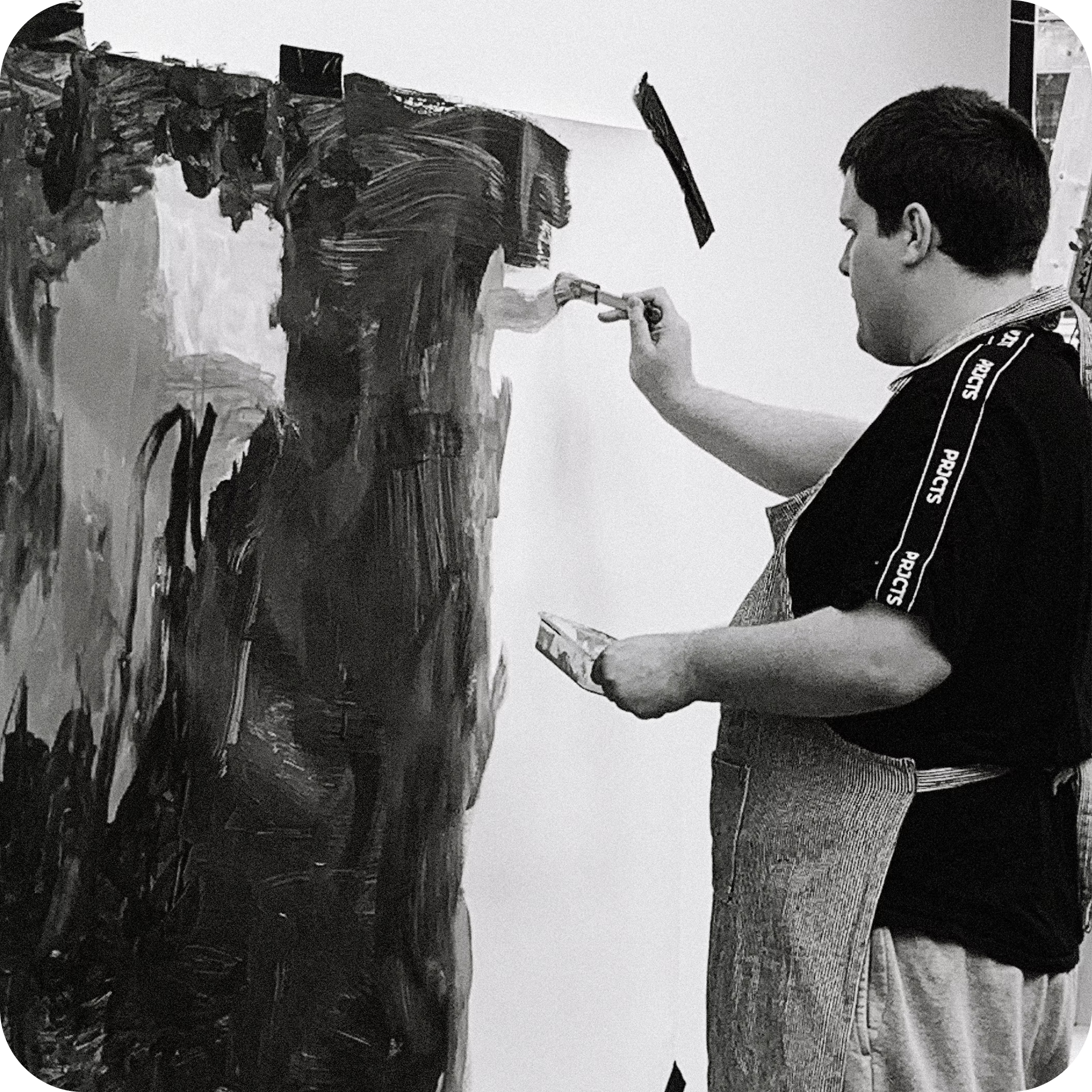 A man with dark hair, wearing a dark T-shirt with text on the sleeve, is painting an abstract street scene on a large canvas in a studio. The artwork features buildings and a street, rendered in bold, expressive brushstrokes.