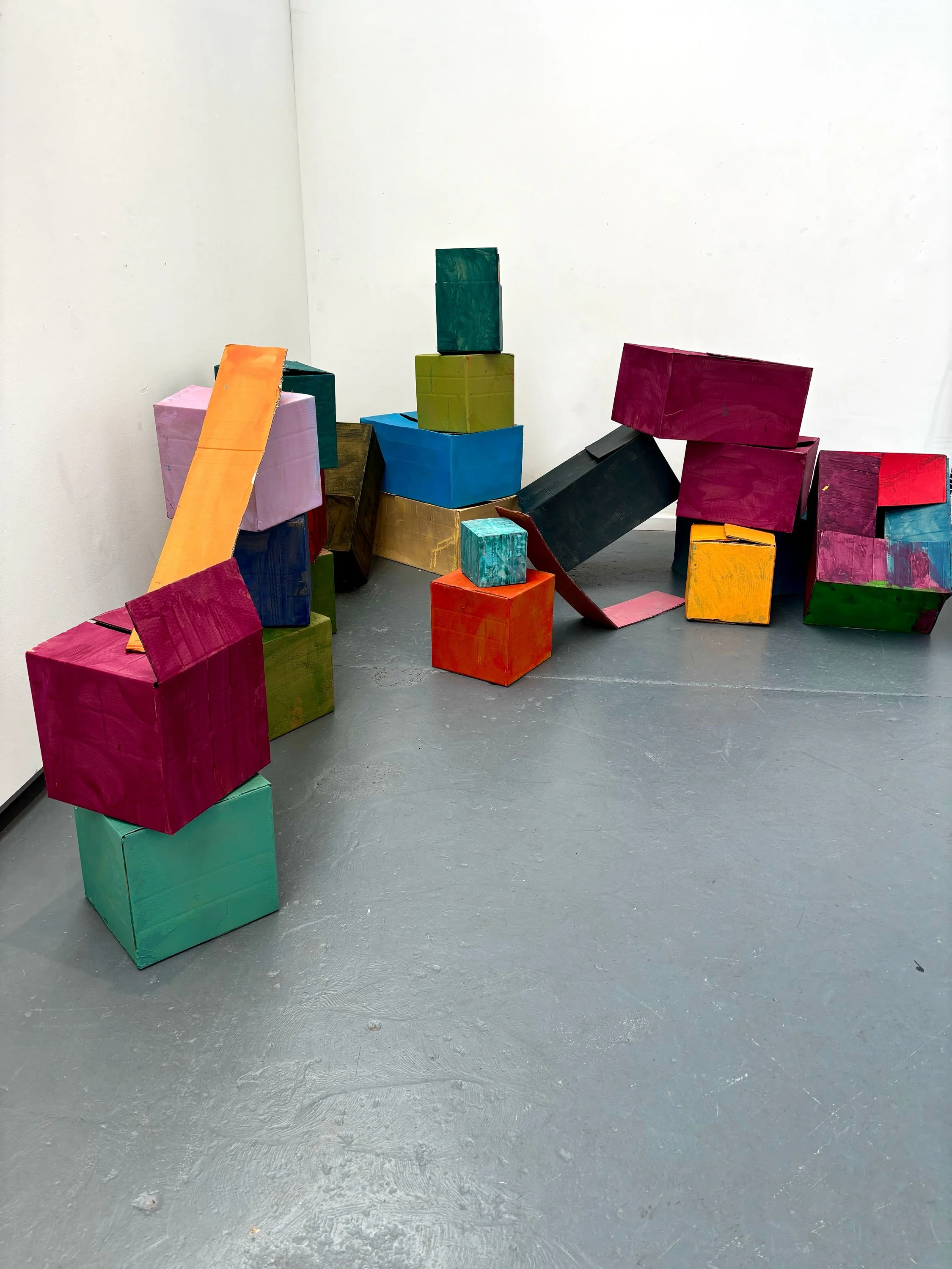 Colorful wooden blocks stacked and scattered on a gray floor against a white wall, forming an abstract sculpture.