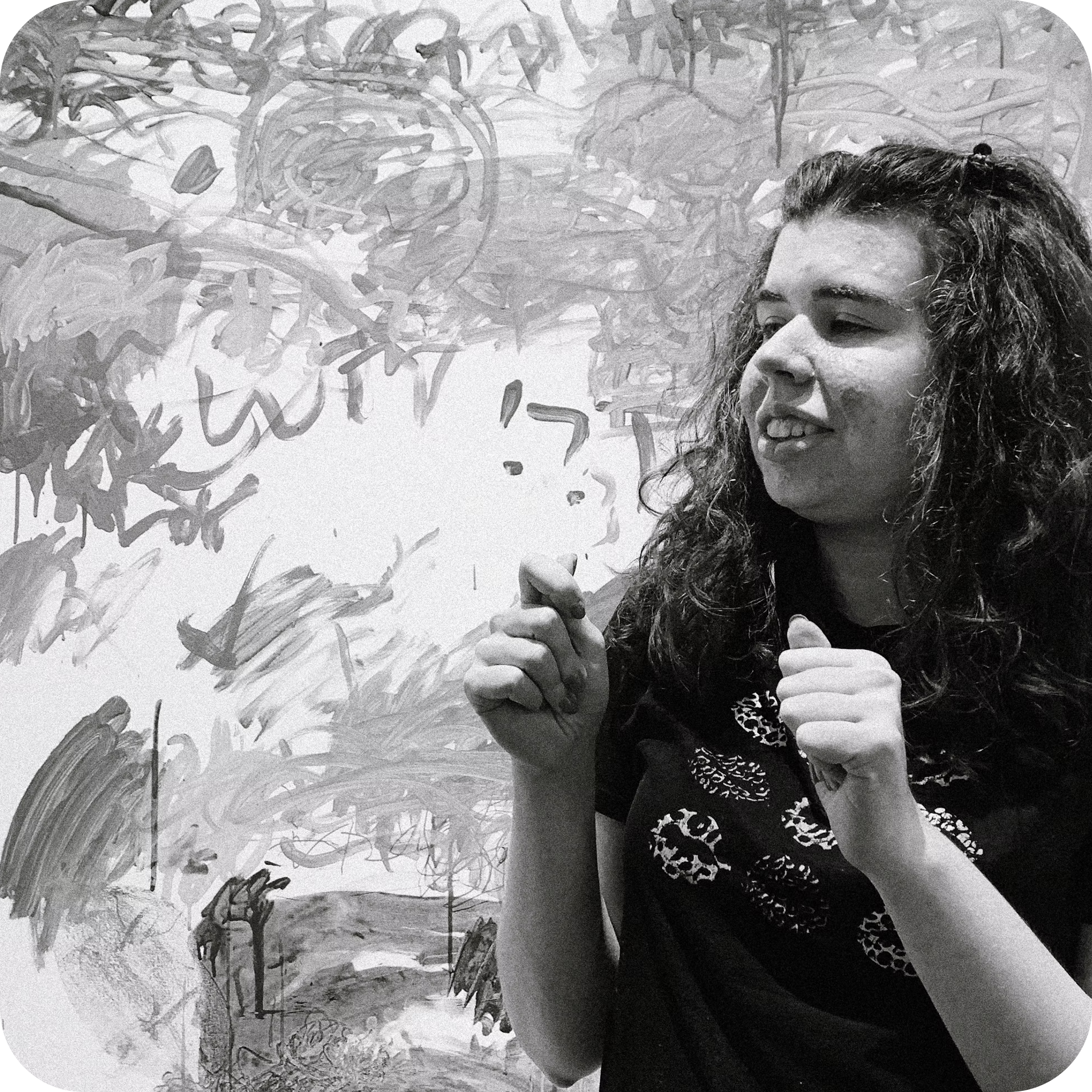 A woman with curly hair and makeup, smiling and pointing forward, standing outdoors near trees with a background of graffiti wall.