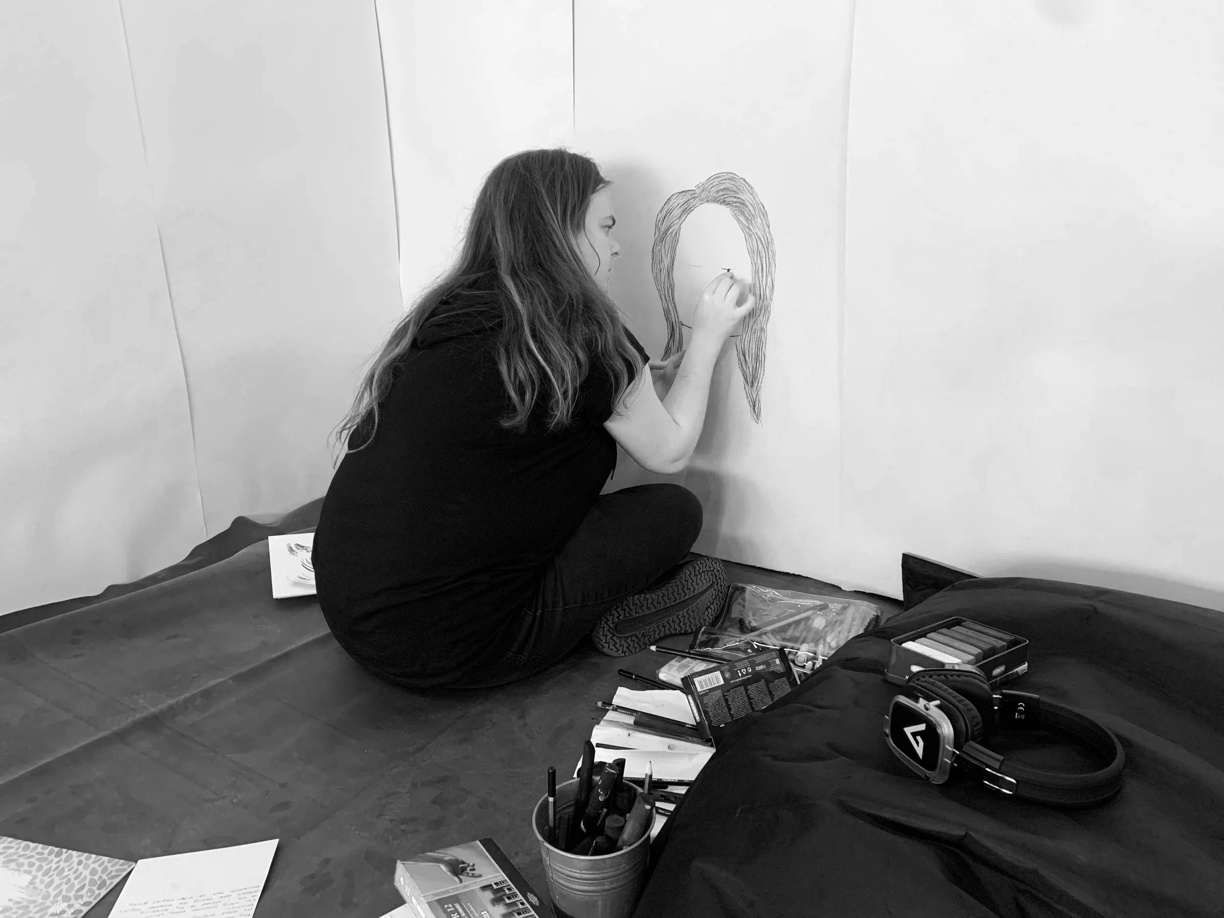 A woman with long hair sitting on the floor, drawing a face on a large sheet of paper attached to a wall. She is surrounded by art supplies, including colored pencils and markers, on the floor and in containers.