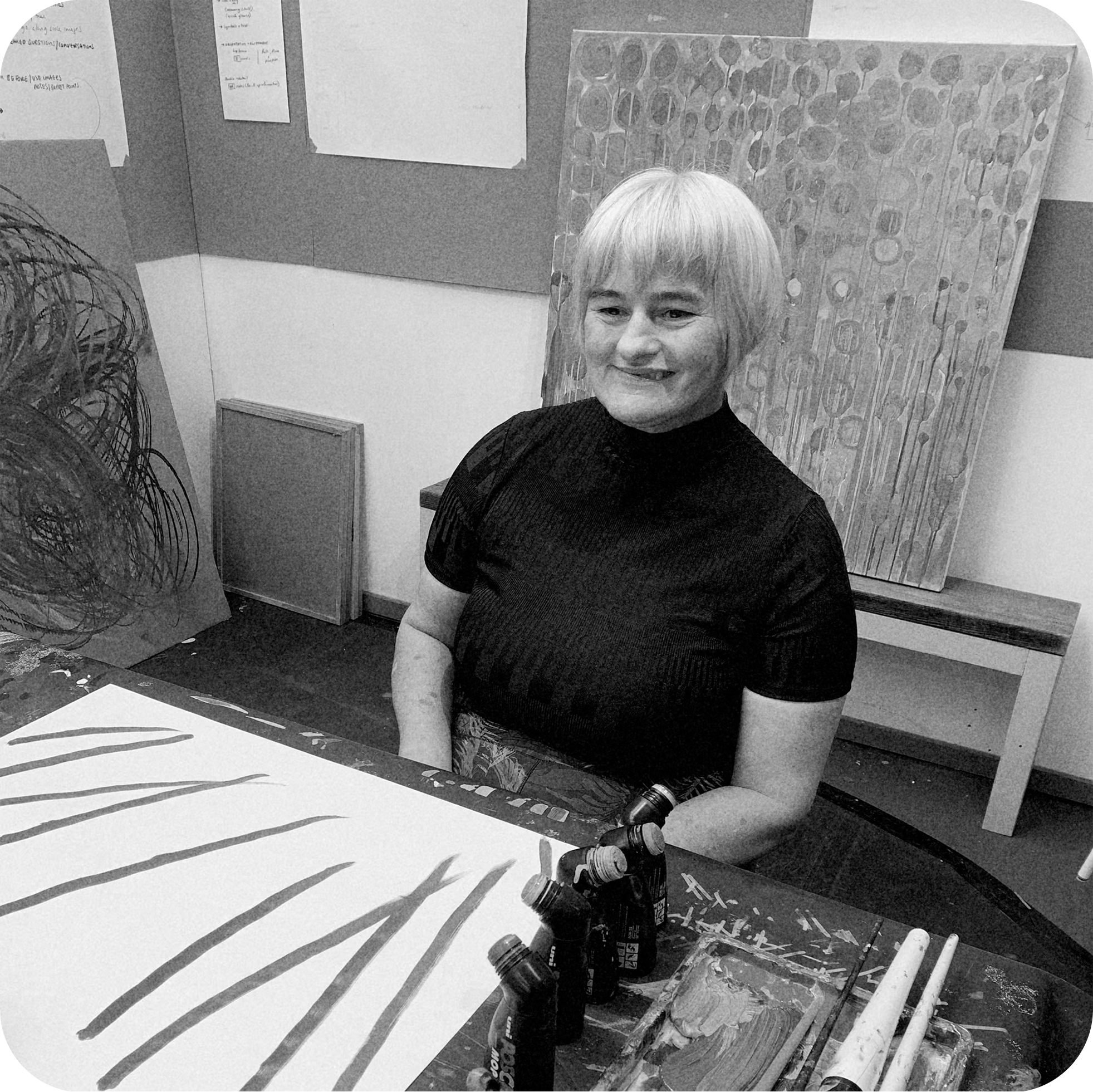 A woman with short blonde hair smiling in an art studio. She is wearing a black top, and there are art supplies including paint bottles, brushes, and papers on the table. In the background, there are abstract paintings and notes pinned on a corkboard.