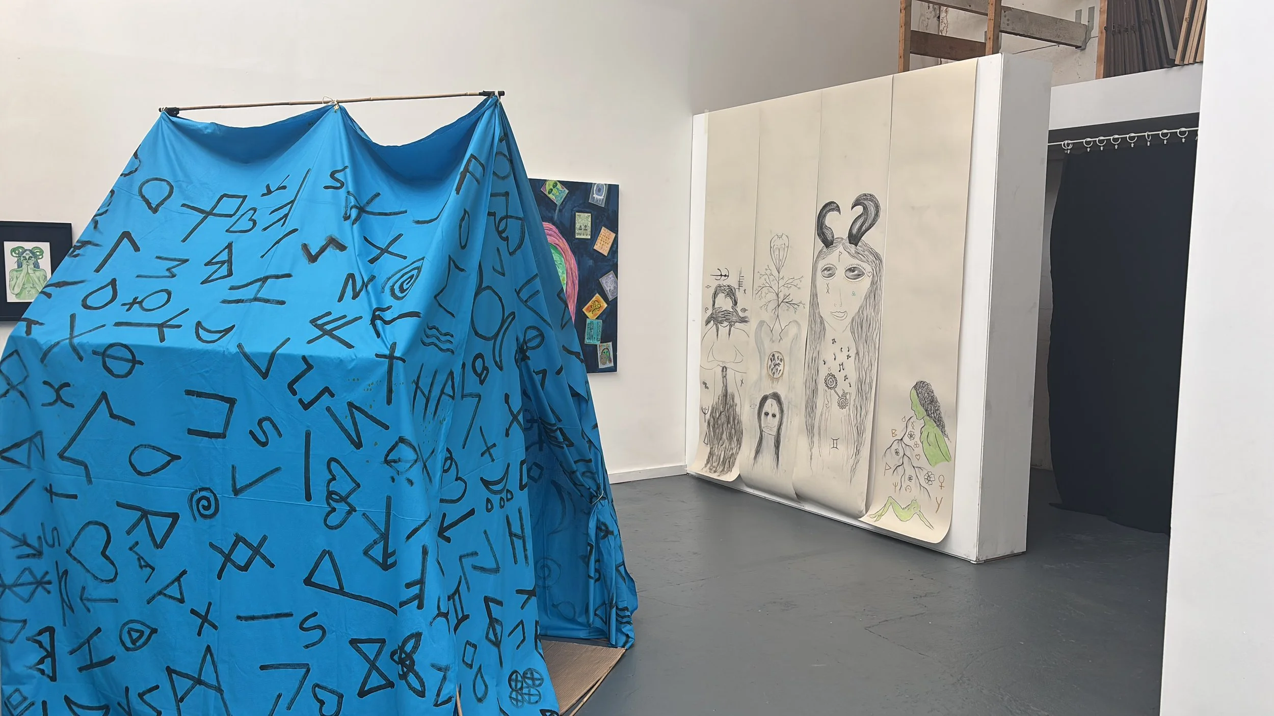 An art gallery with a large blue fabric covered object decorated with black abstract symbols, and a sheet of paper with various sketches of faces and symbols leaning against the wall.