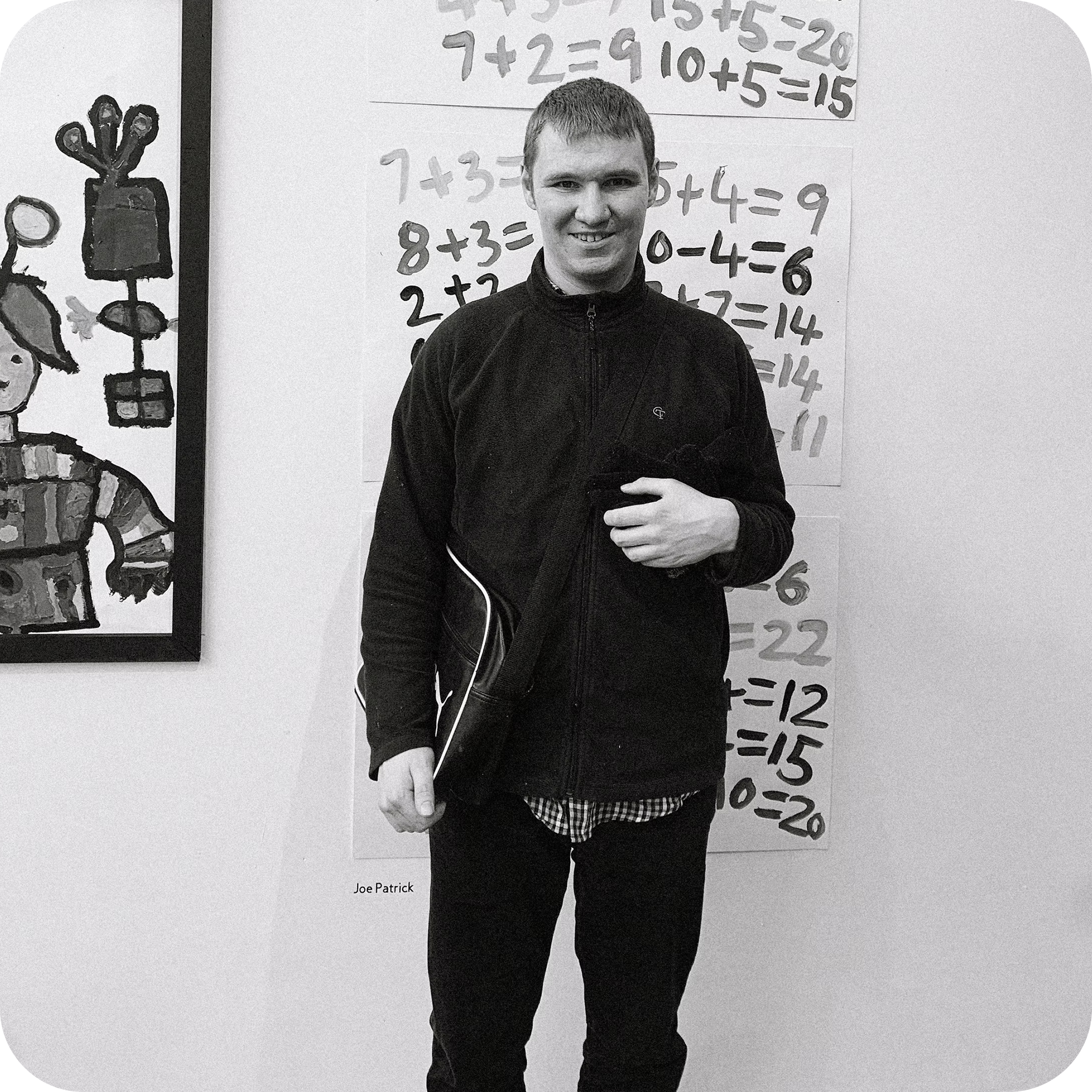 A young man with short hair and a smile, wearing a black jacket, standing in front of a white wall with math problems written on a sheet of paper attached to the wall. To his left, there is a painted cartoon-like figure of a creature with multiple arms.