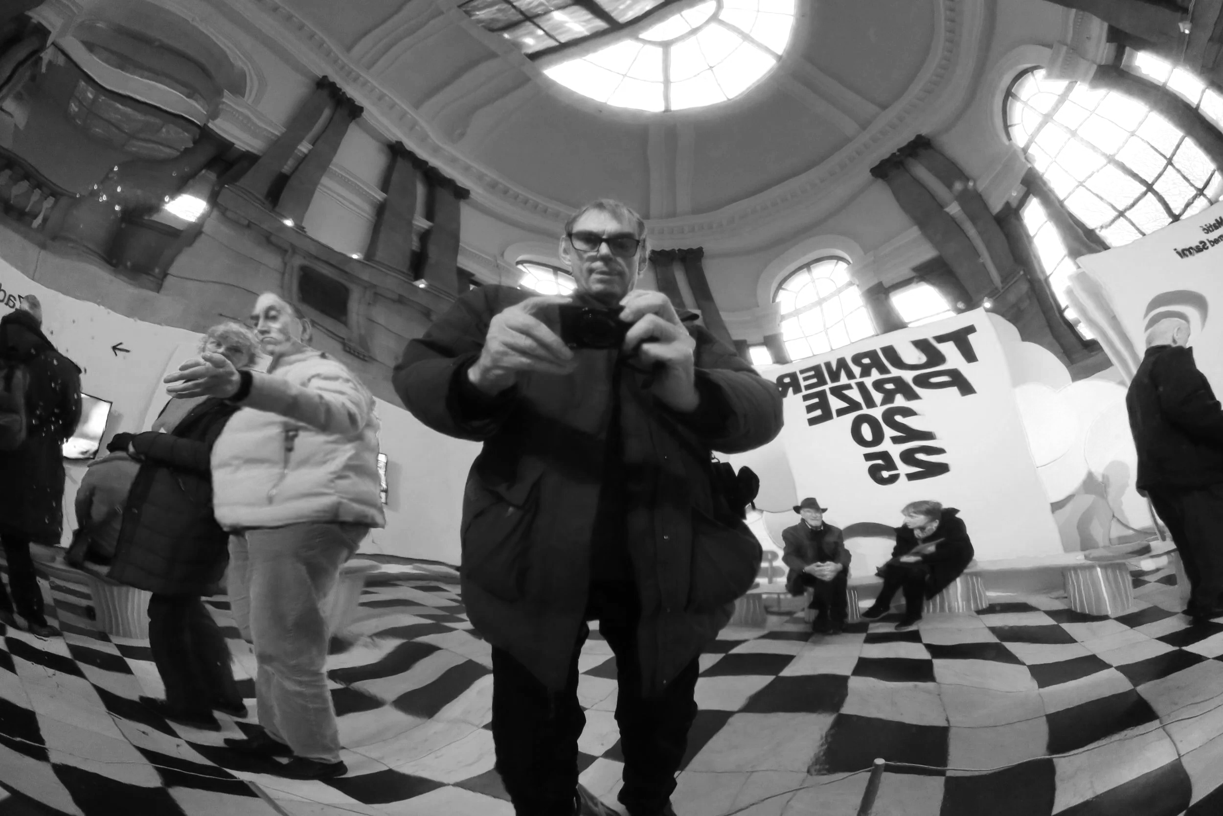A man with glasses is taking a photo of himself using a large convex mirror in a grand indoor space with ornate architecture, high ceilings with a large skylight, and multiple other people around him. Some individuals are looking at their phones, and there are banners with text in the background.