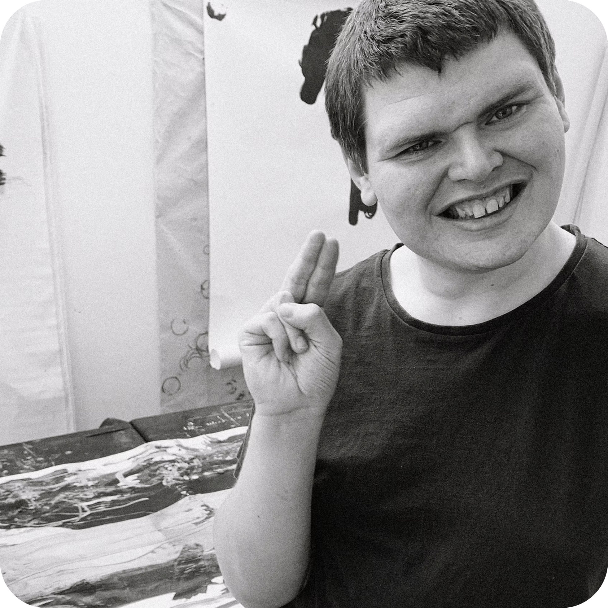 Black and white photo of a smiling young man with short, wavy hair, showing his teeth, holding up his right hand with fingers spread, and making a peace sign. Behind him, part of a poster or painting with abstract shapes and a landscape scene with water, sky, and some trees.