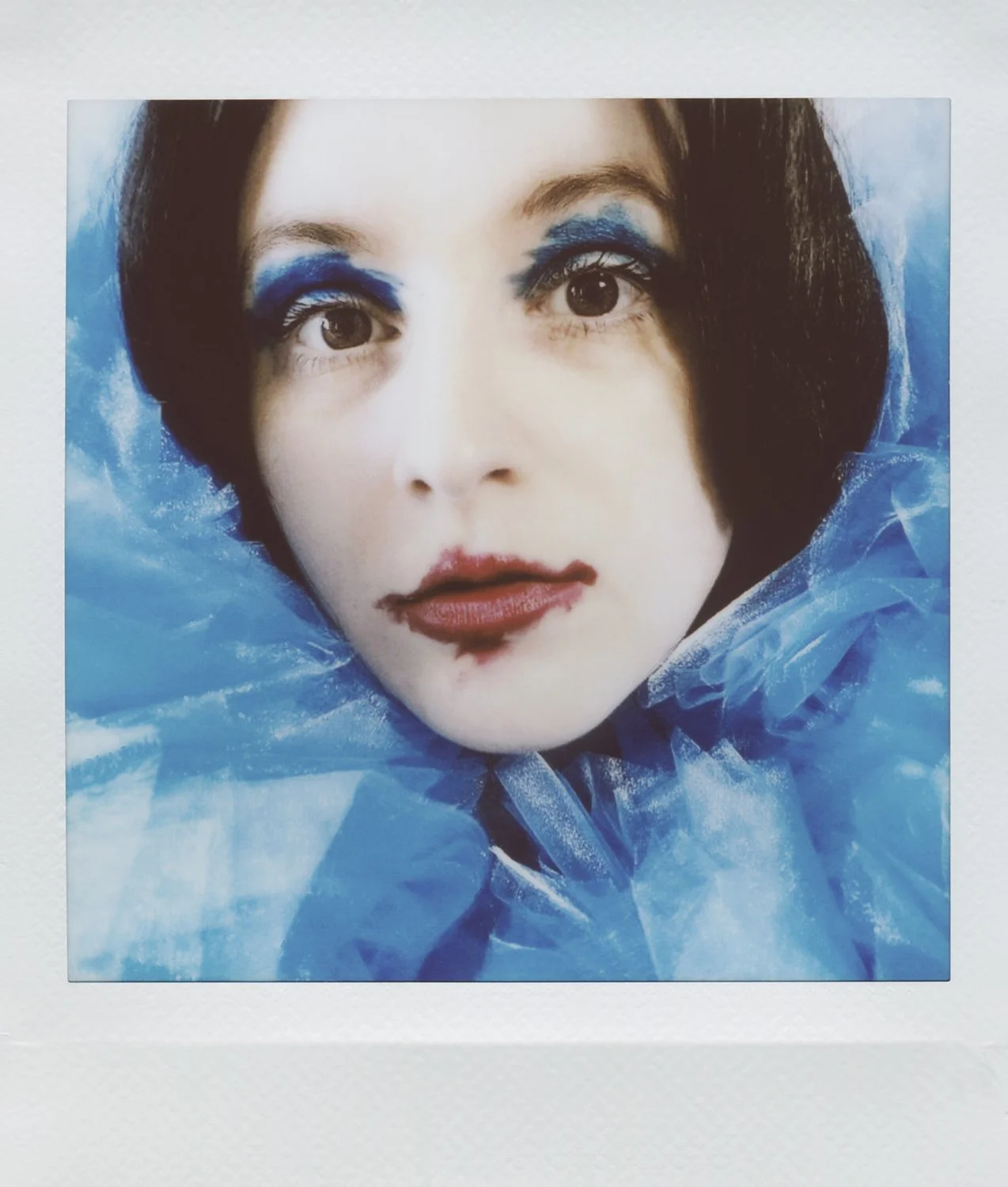 Close-up of a woman's face with dramatic blue eyeshadow and lipstick, surrounded by blue tulle fabric.