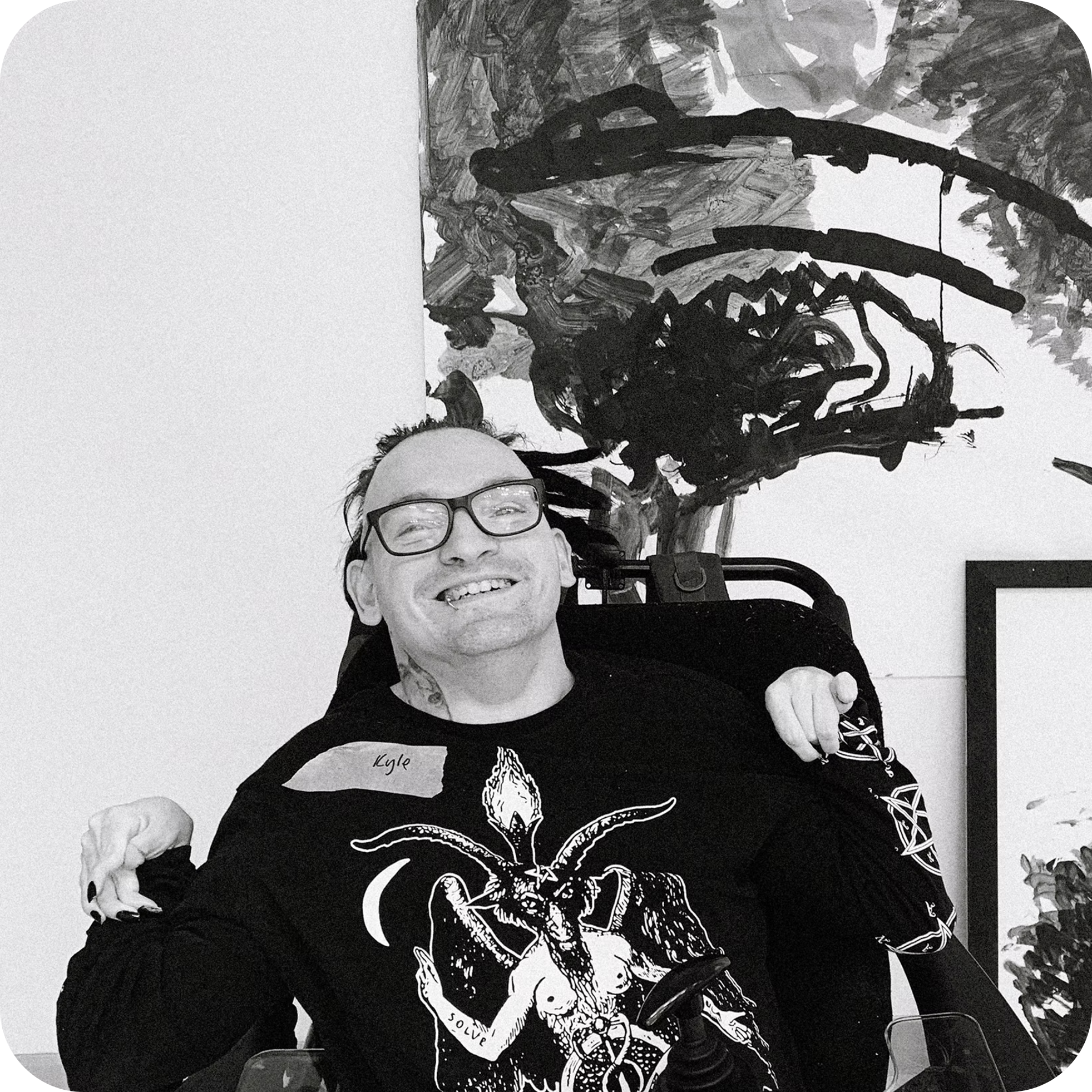 A person with glasses smiling, wearing a sweater with a large graphic of a demonic creature with horns and wings print on it, sitting at a table with empty wine glasses, with a black and white abstract painting behind them.