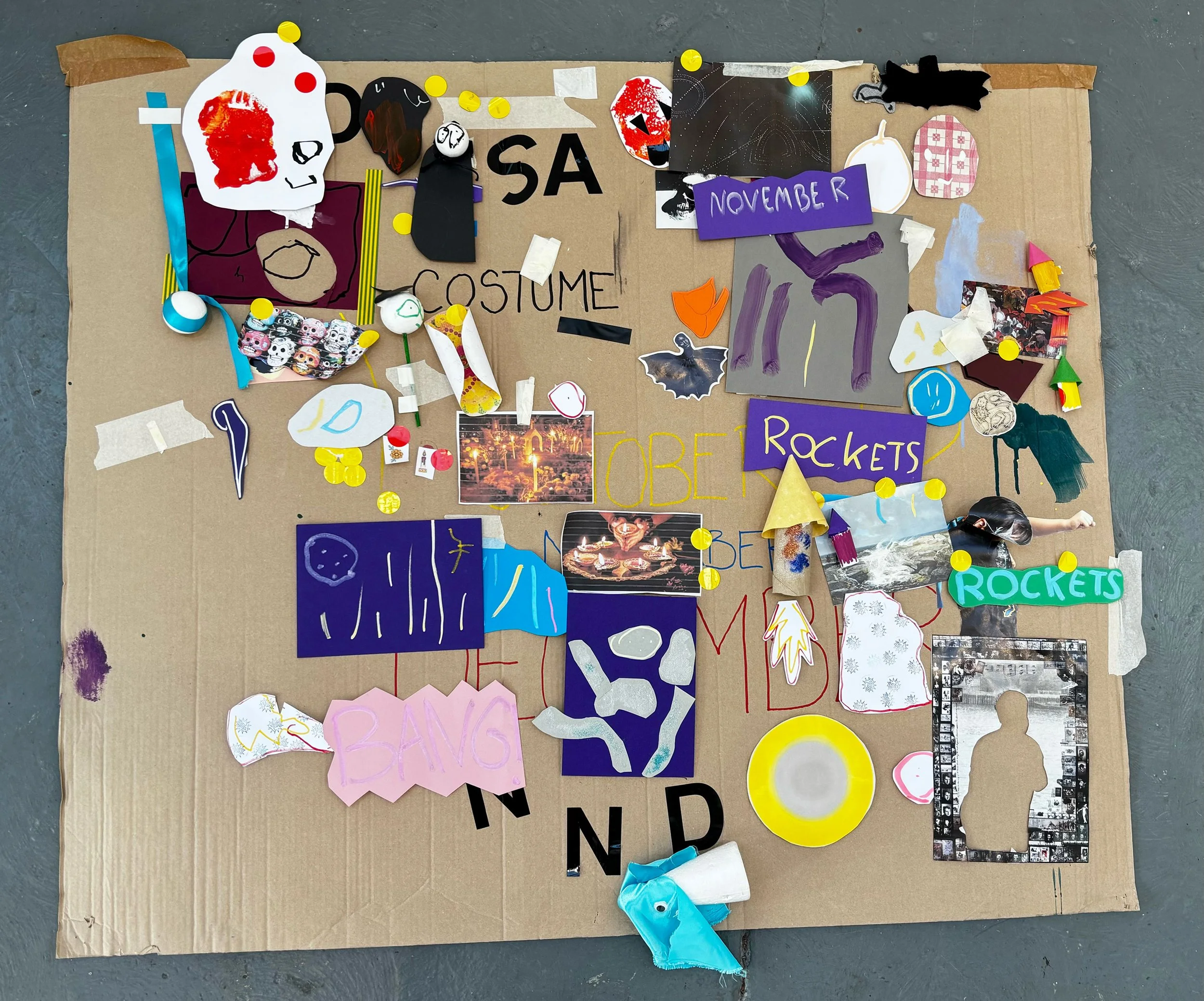 A large piece of cardboard with various handmade decorations, drawings, and signs attached to it. The decorations include paper cutouts, drawings, painted objects, and photographs. Some signs display words such as 'COSTUME', 'NOVEMBER', 'ROCKETS', an