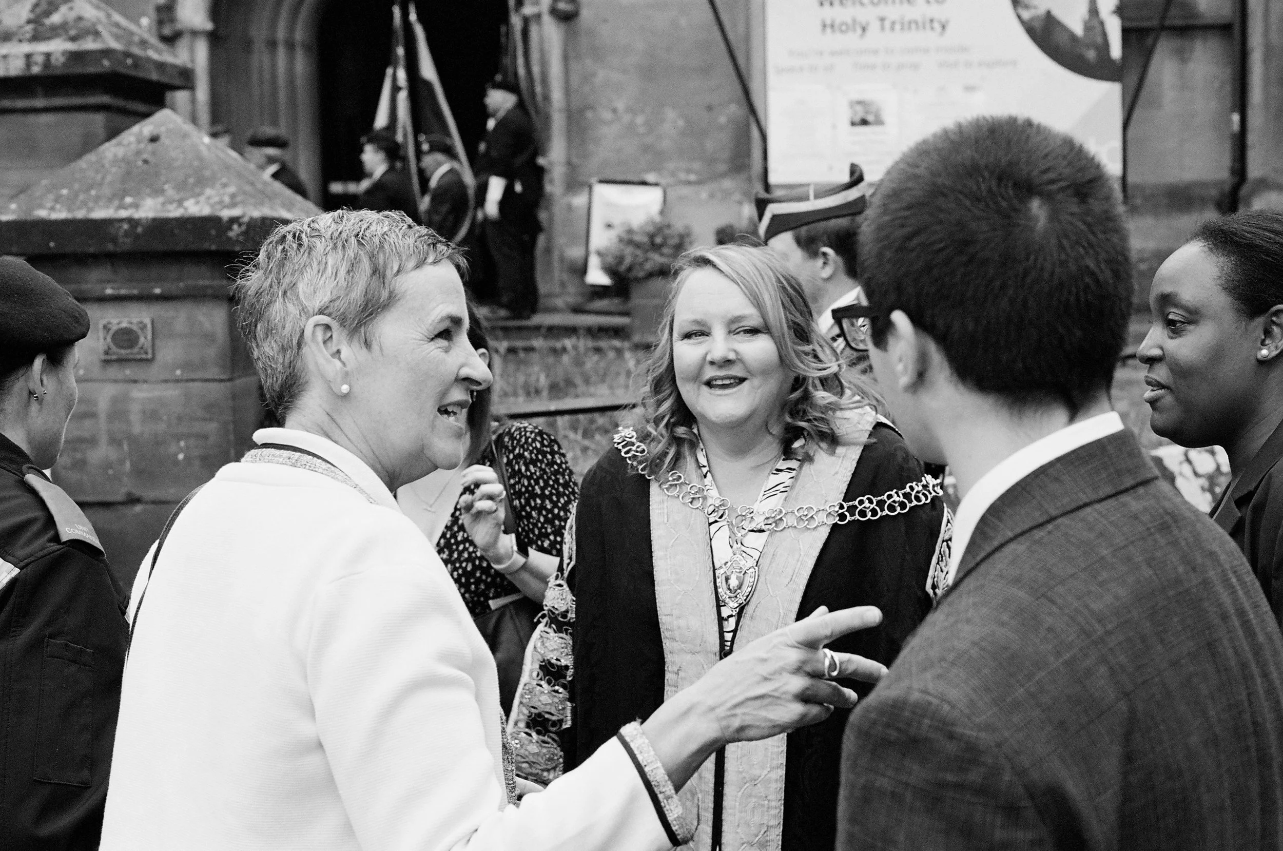 Black and white photo of a group of people in formal attire, engaged in conversation outdoors, with a church or similar building in the background.