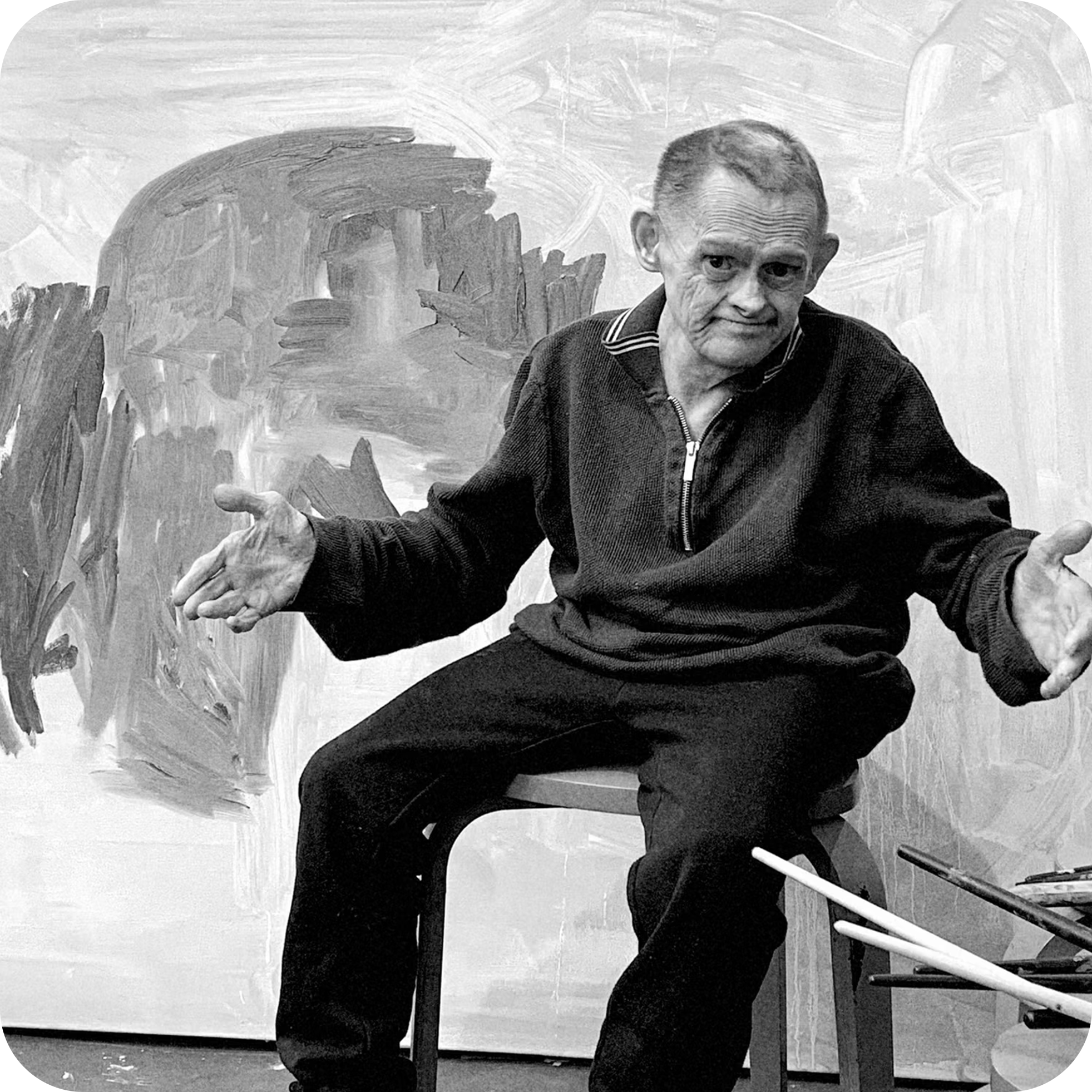 A black and white photo of a middle-aged man with short hair, wearing a zip-up sweater, sitting at a table in front of a painted abstract background. He appears to be gesturing with his right hand, and there are a few chairs or objects partly visible on the right side of the image.