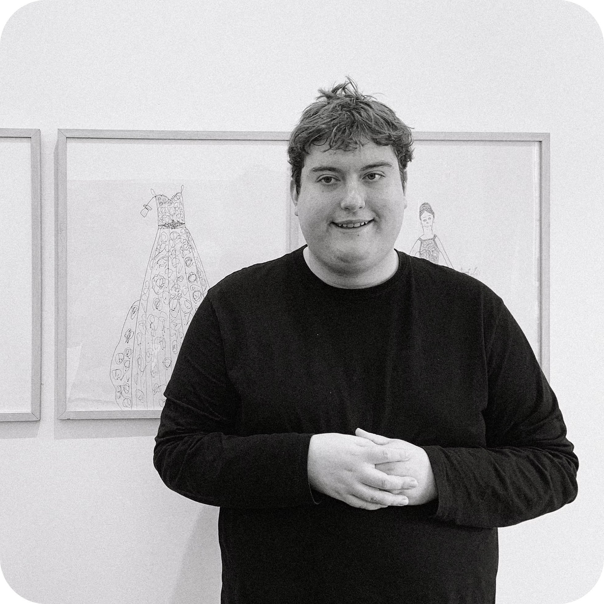 A young man with curly hair and a friendly smile is standing in front of artwork on a gallery wall. He is wearing a black long-sleeve shirt and has his hands crossed over his stomach.