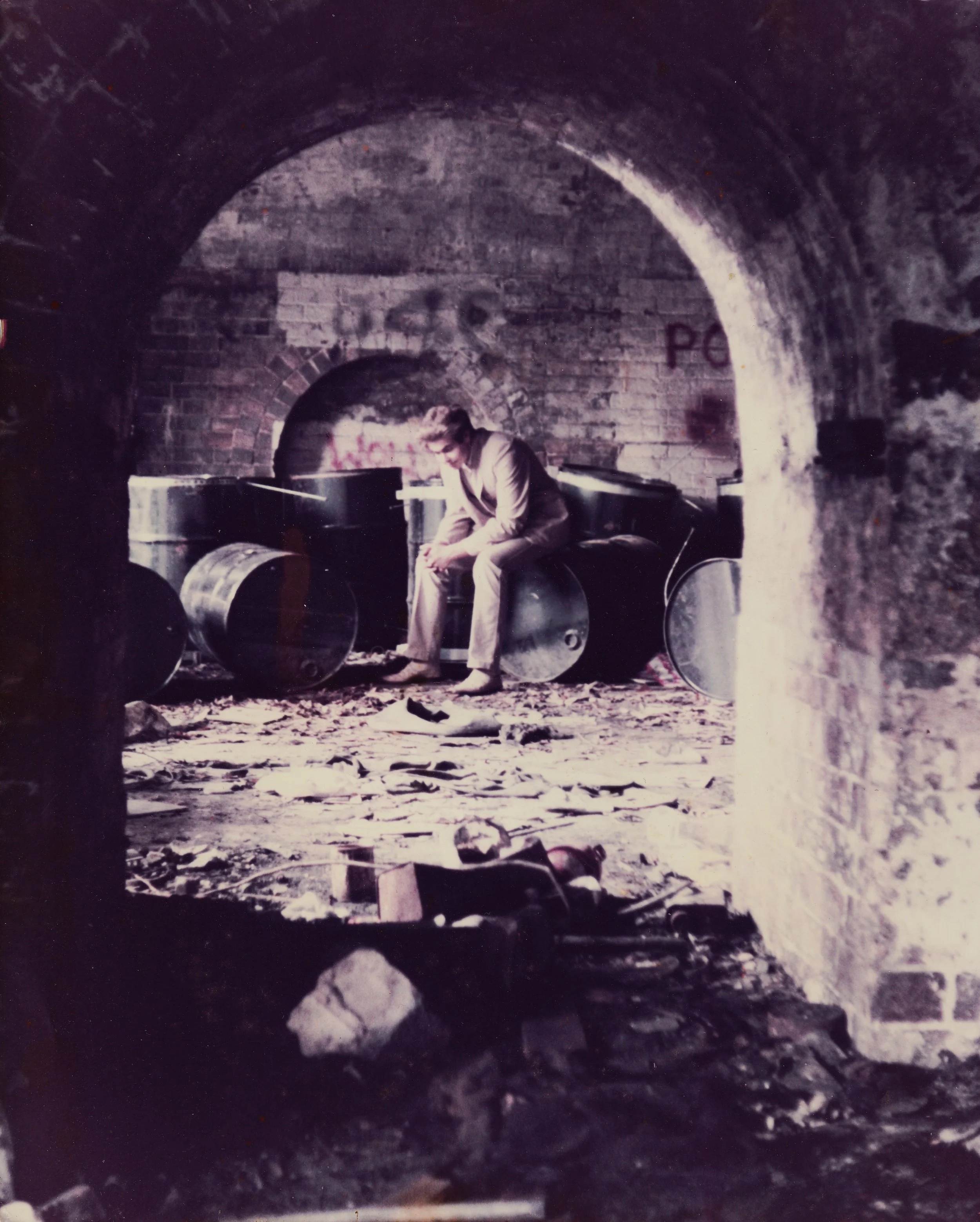 A person sitting on a stool in front of a brick arch, in a dimly lit underground tunnel with barrels and graffiti.