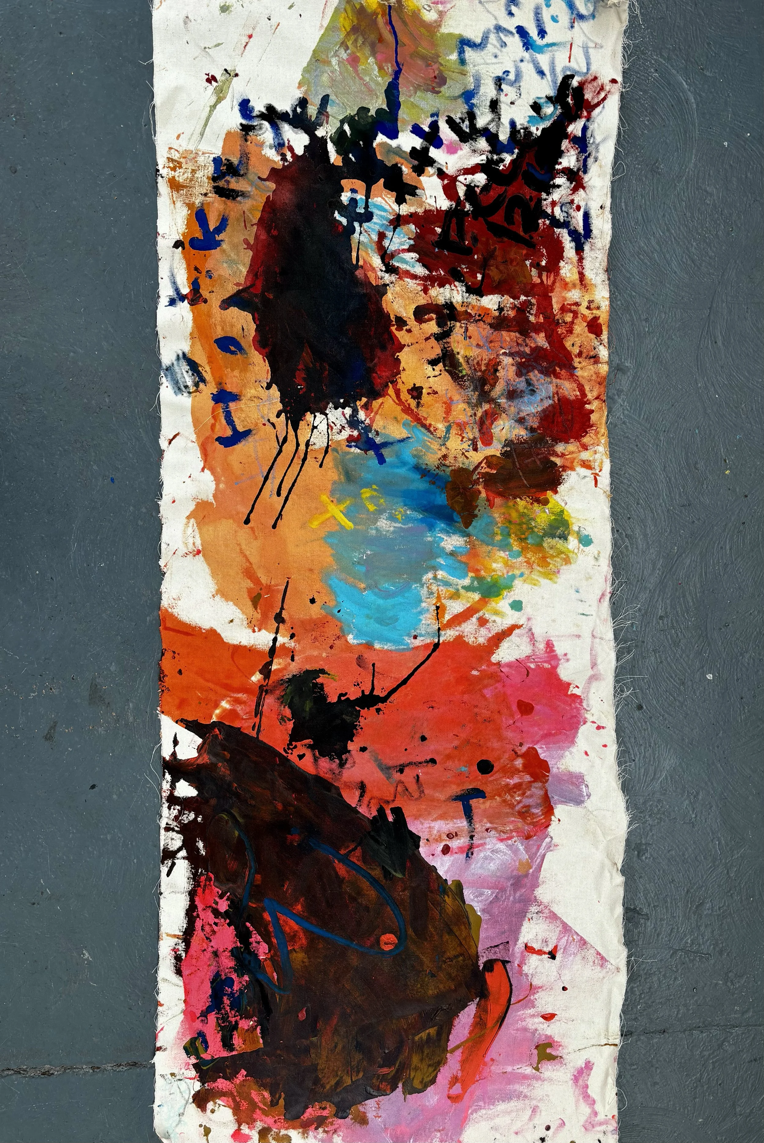 Abstract graffiti artwork on a vertical strip of paper or fabric, featuring bold black, red, orange, blue, pink, and yellow splashes, strokes, and scribbles, mounted on a gray textured surface.