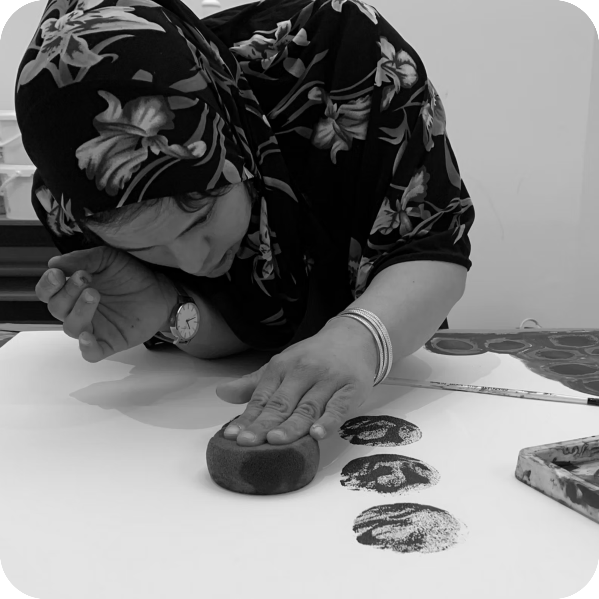 Close-up of a person's arm with a floral-patterned shirt, wearing a wristwatch and bracelets, resting on a white surface with rocks and stamped footprints, in black and white.