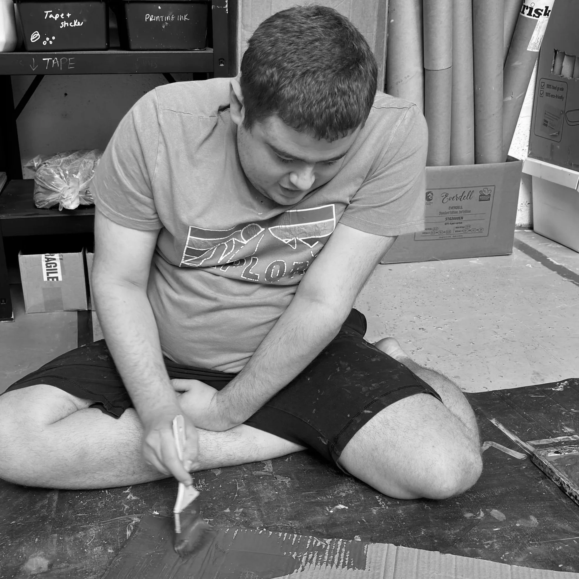 A man sitting on the floor with his head bowed down and eyes closed, surrounded by boxes and shelves filled with tape, cardboard, and labels, in what appears to be a storage or warehouse area.