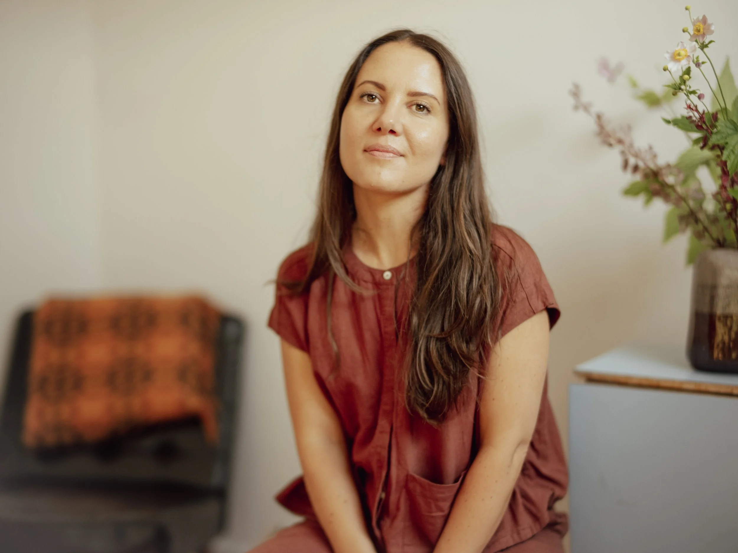A woman with long brown hair and light skin wearing a rust-colored top sitting on a chair in a room with light-colored walls and flowers.