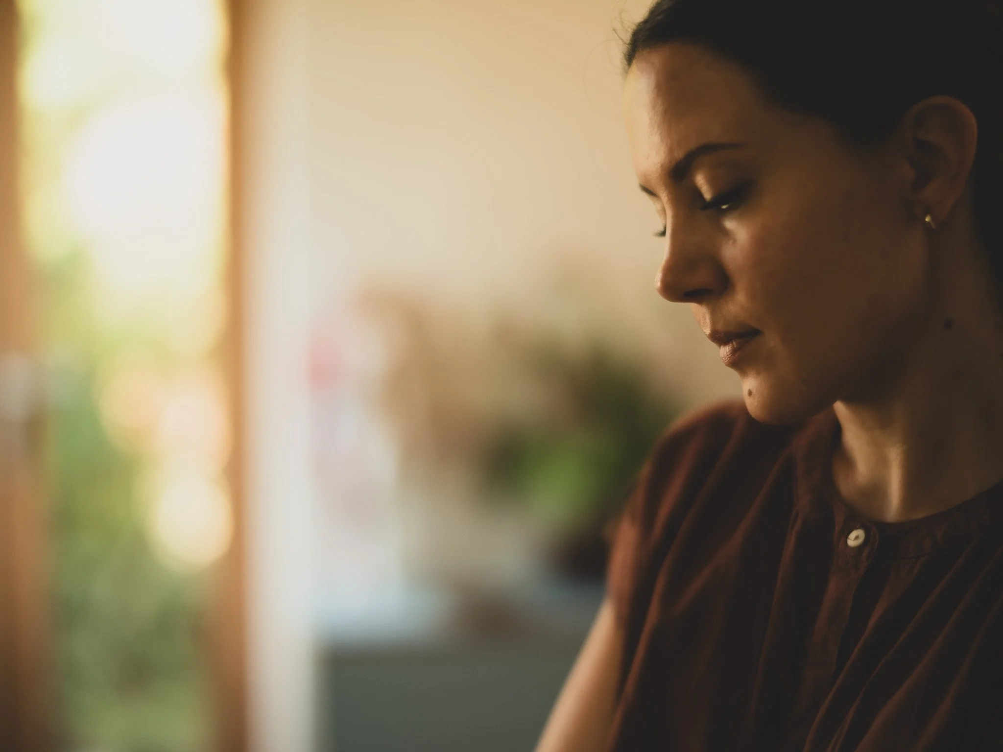 Close-up of a woman with short dark hair and earrings, looking downward with a contemplative expression, softly lit with a blurred background.