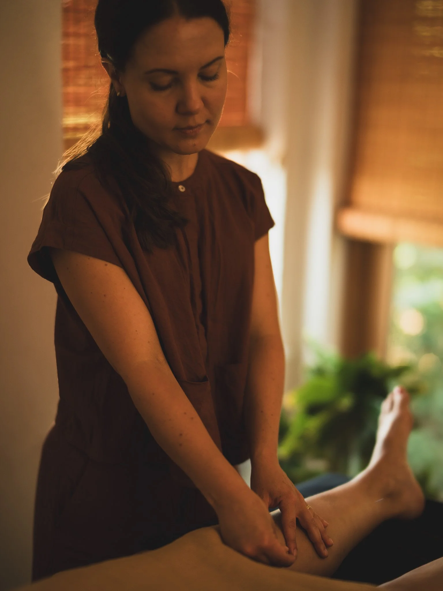 A woman is giving a massage to a person lying down, with her hands on their legs or feet. The setting appears to be indoors with warm lighting and a background of window blinds and plants.