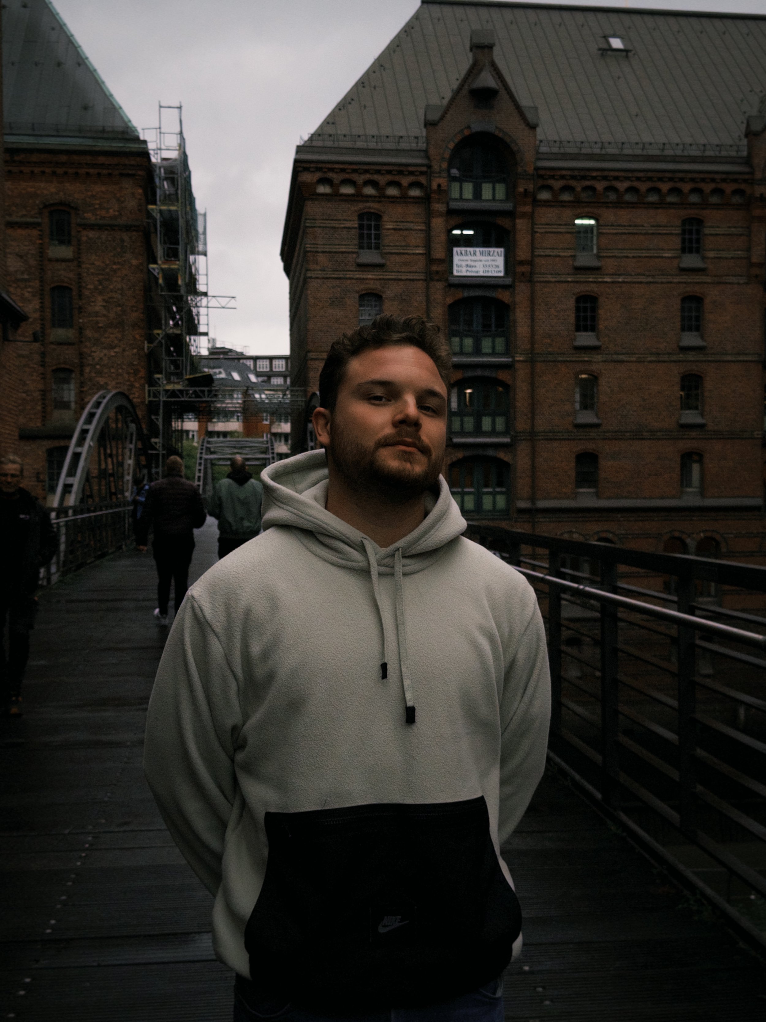 A young man with a beard and curly hair standing on a bridge during overcast weather, wearing a light-colored hoodie with a black Nike logo pocket, with historic brick buildings in the background.