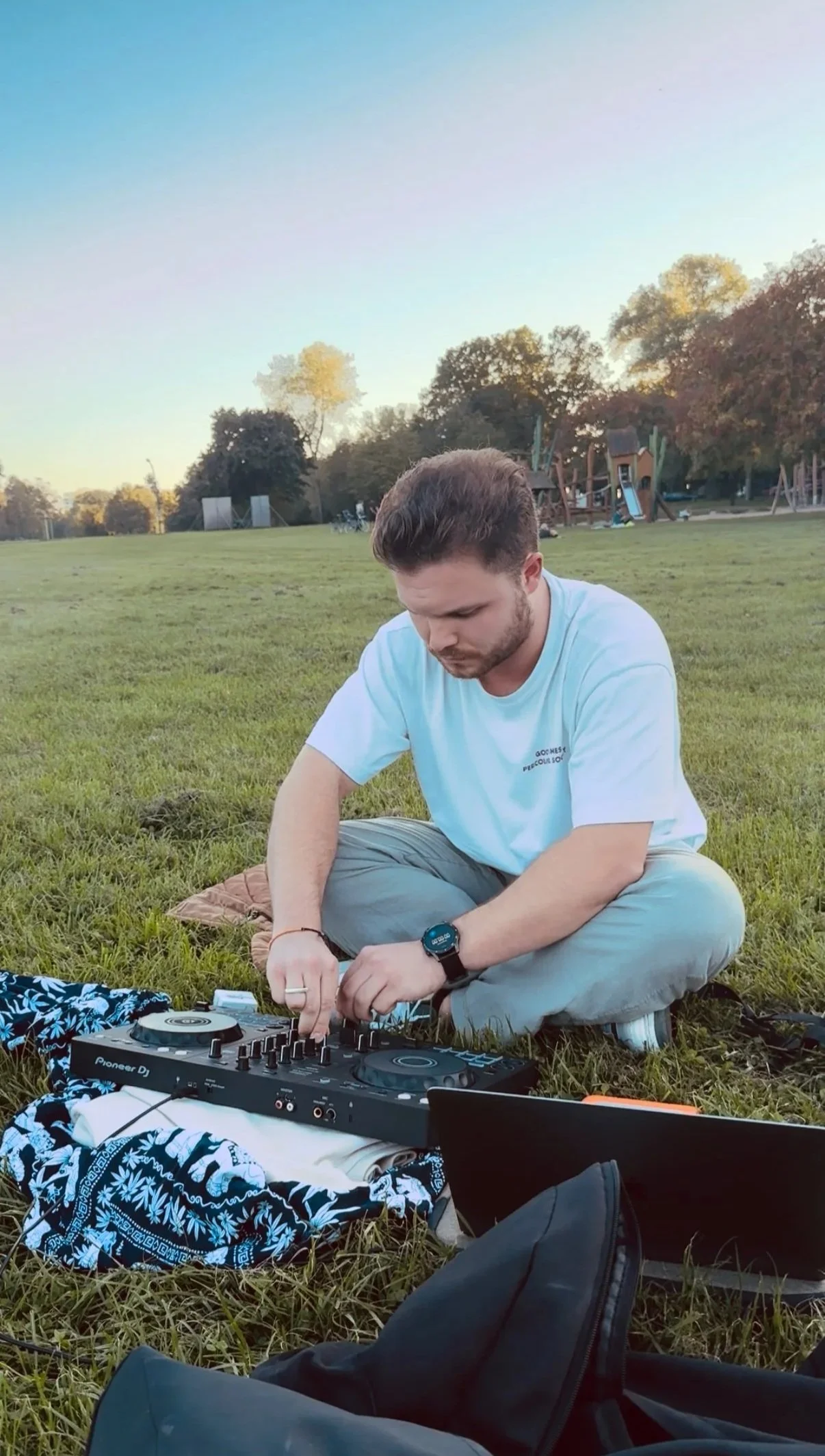 A man sitting cross-legged on the grass using a DJ mixer and laptop outdoors at sunset.