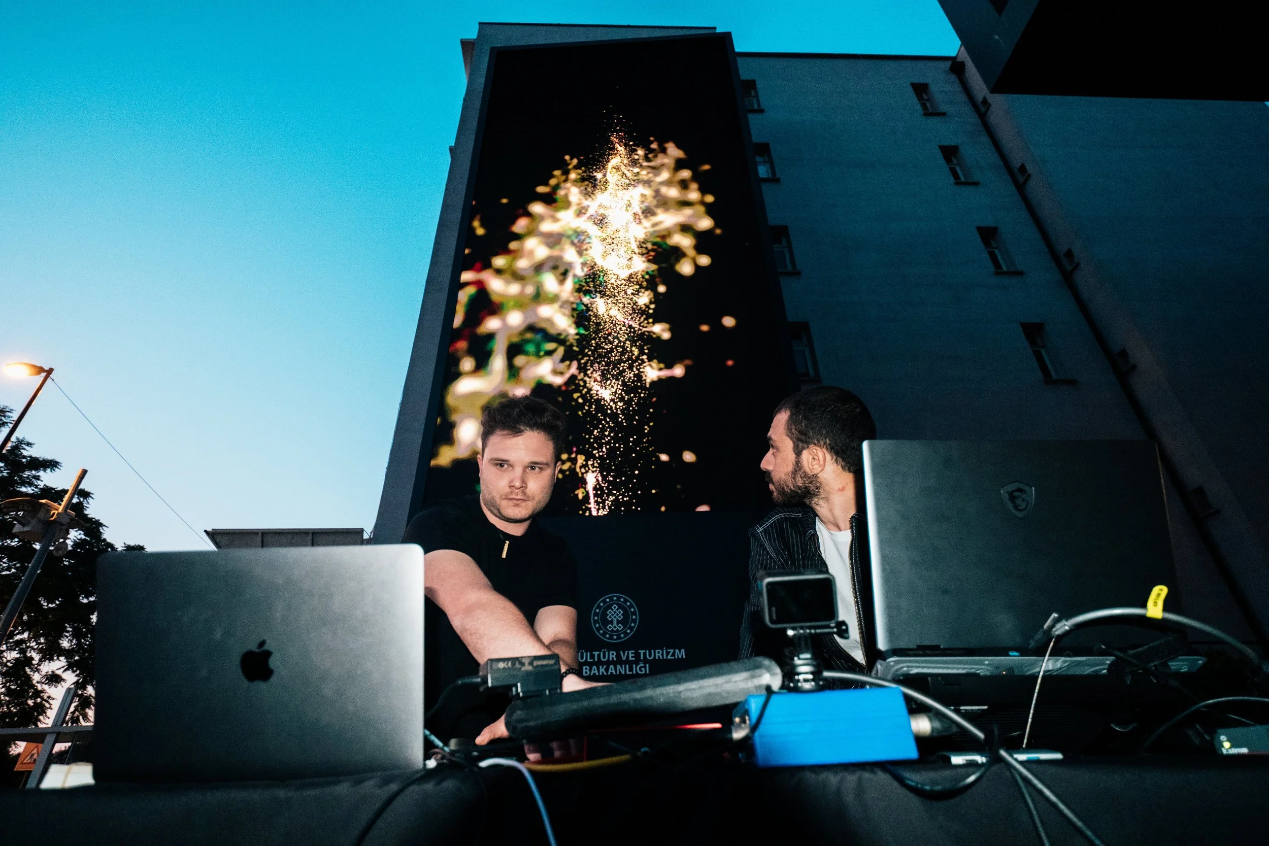 Two men are at a nighttime outdoor event with laptops and equipment on a table. Behind them, there is a large LED display showing visuals against a building in the background.