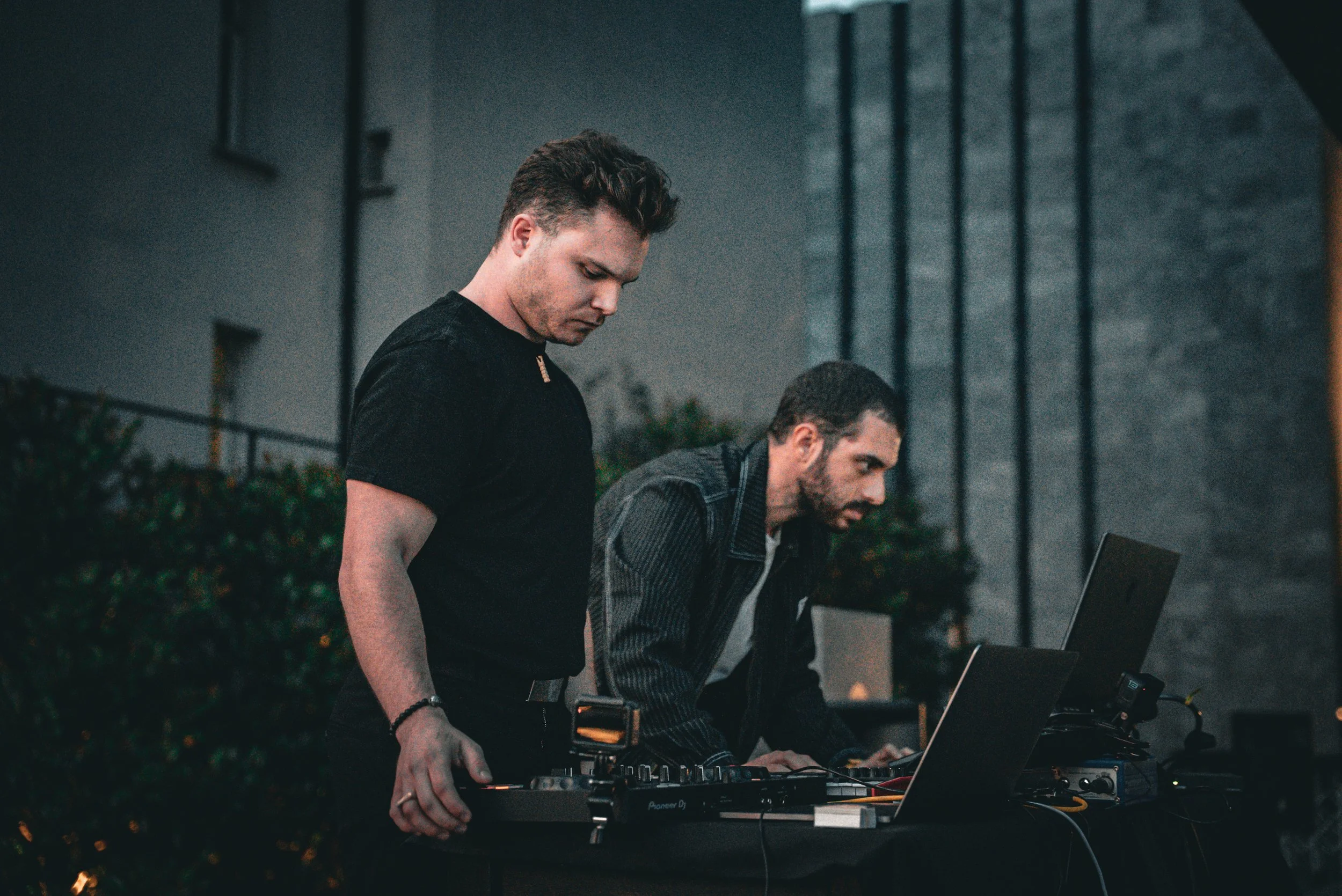 Two men DJing outdoors at night, using laptops and DJ equipment, with a dark urban setting in the background.