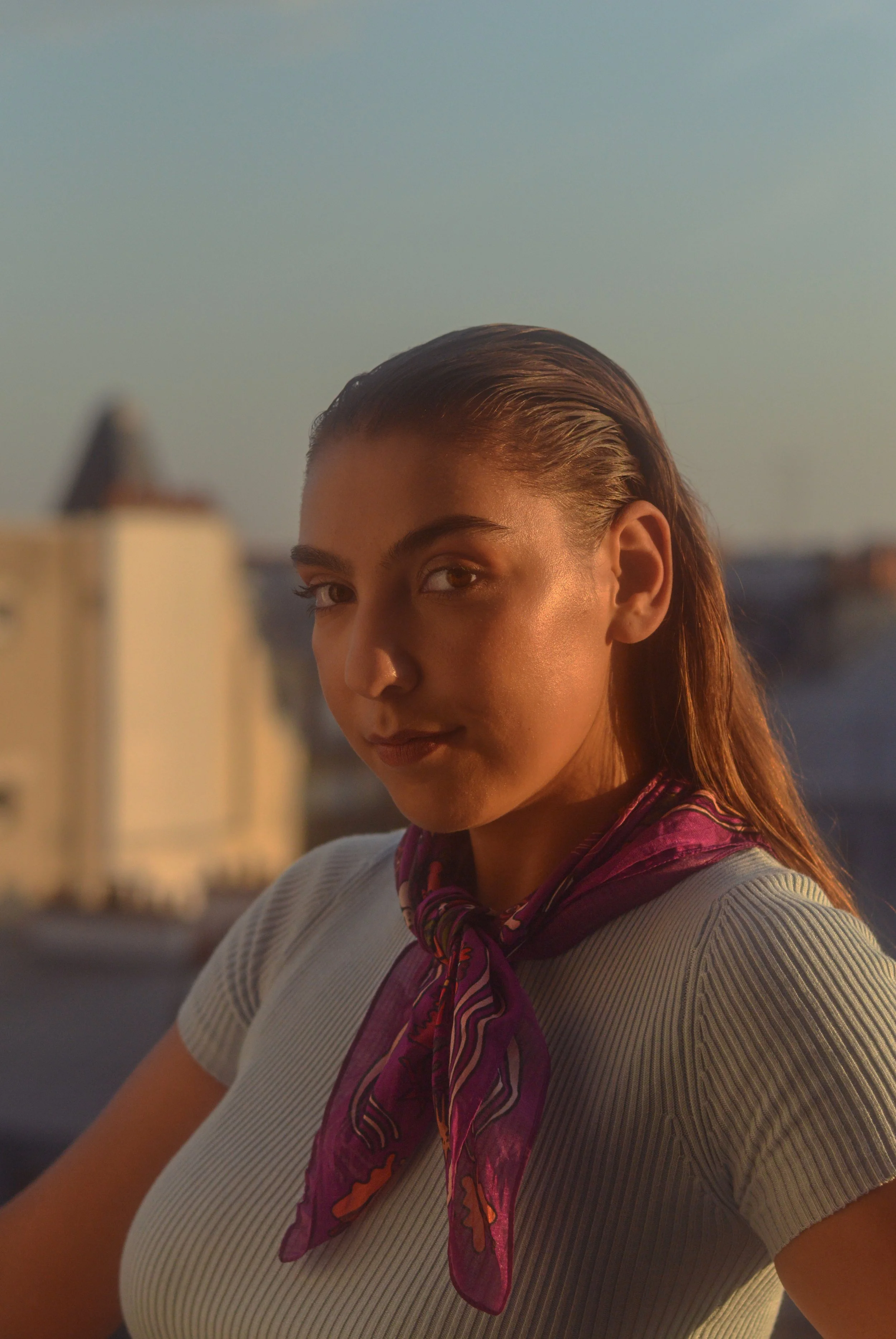 Jeune femme avec cheveux humides, portant un t-shirt rayé blanc et violet, et un foulard violet avec motifs, regardant vers la caméra en plein air lors du coucher de soleil.