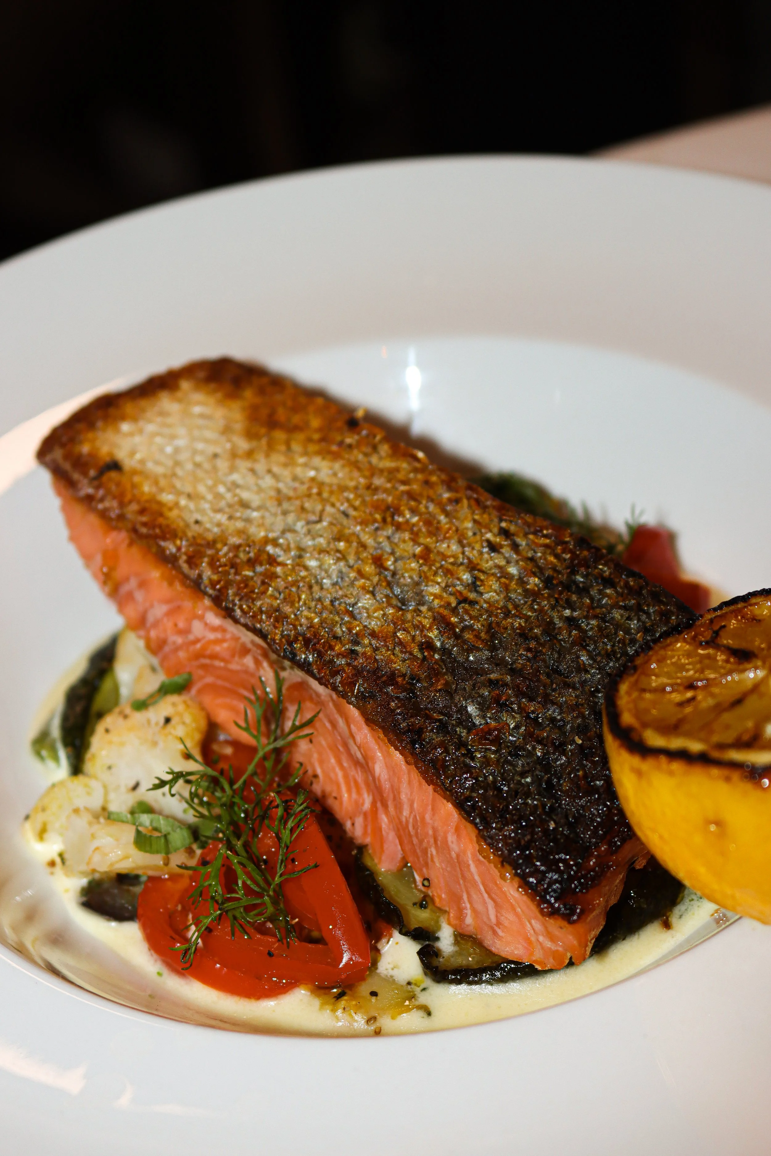 Filet de saumon grillé avec peau croustillante, accompagné de légumes grillés et d'une crème ou sauce en dessous, dans une assiette blanche.