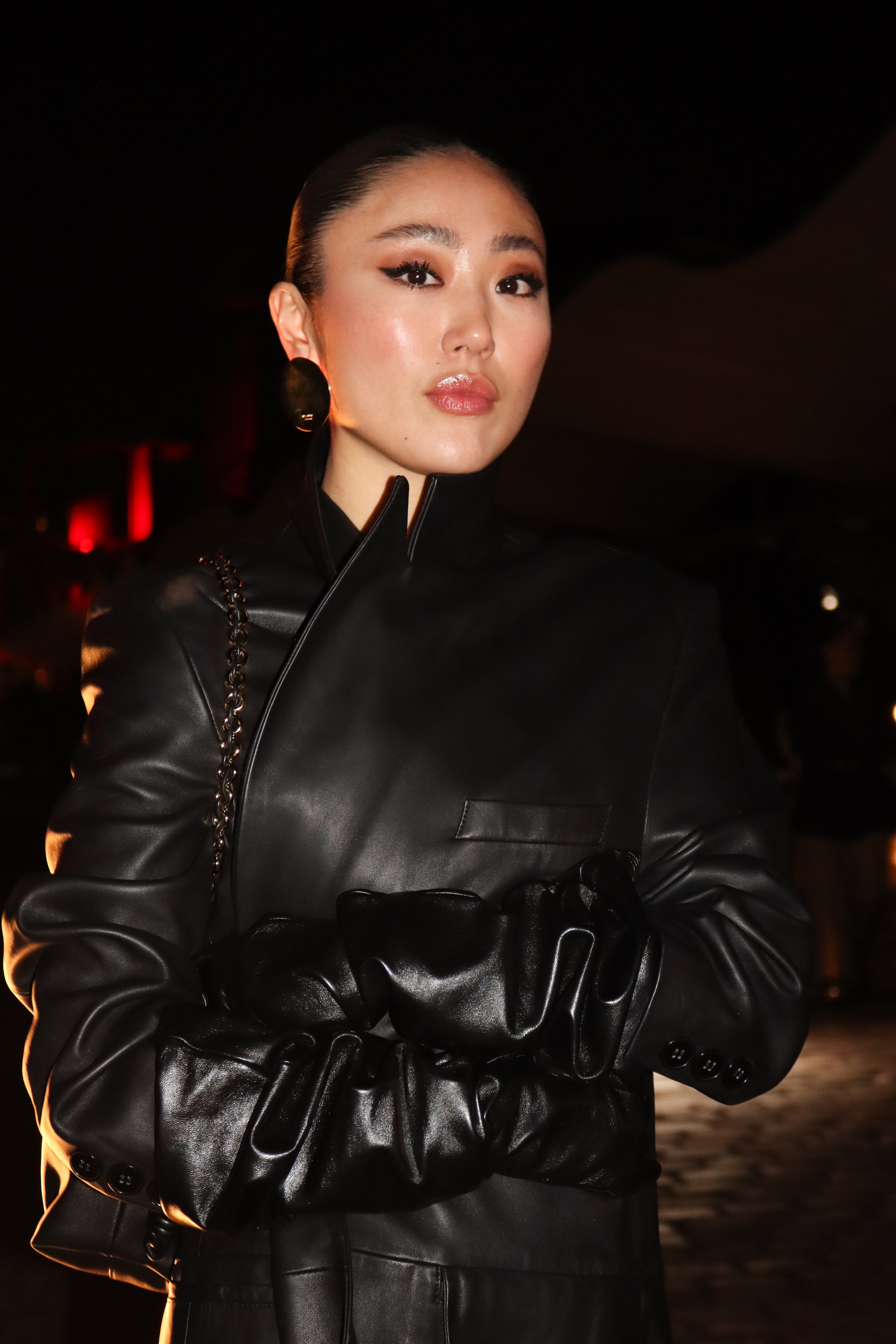 Photographie de portrait: Femme élégante portant un manteau en cuir noir avec gants et boucles d'oreilles rondes dans un environnement sombre avec éclairage chaud. Personnalité invité à la Fashion week de Paris.