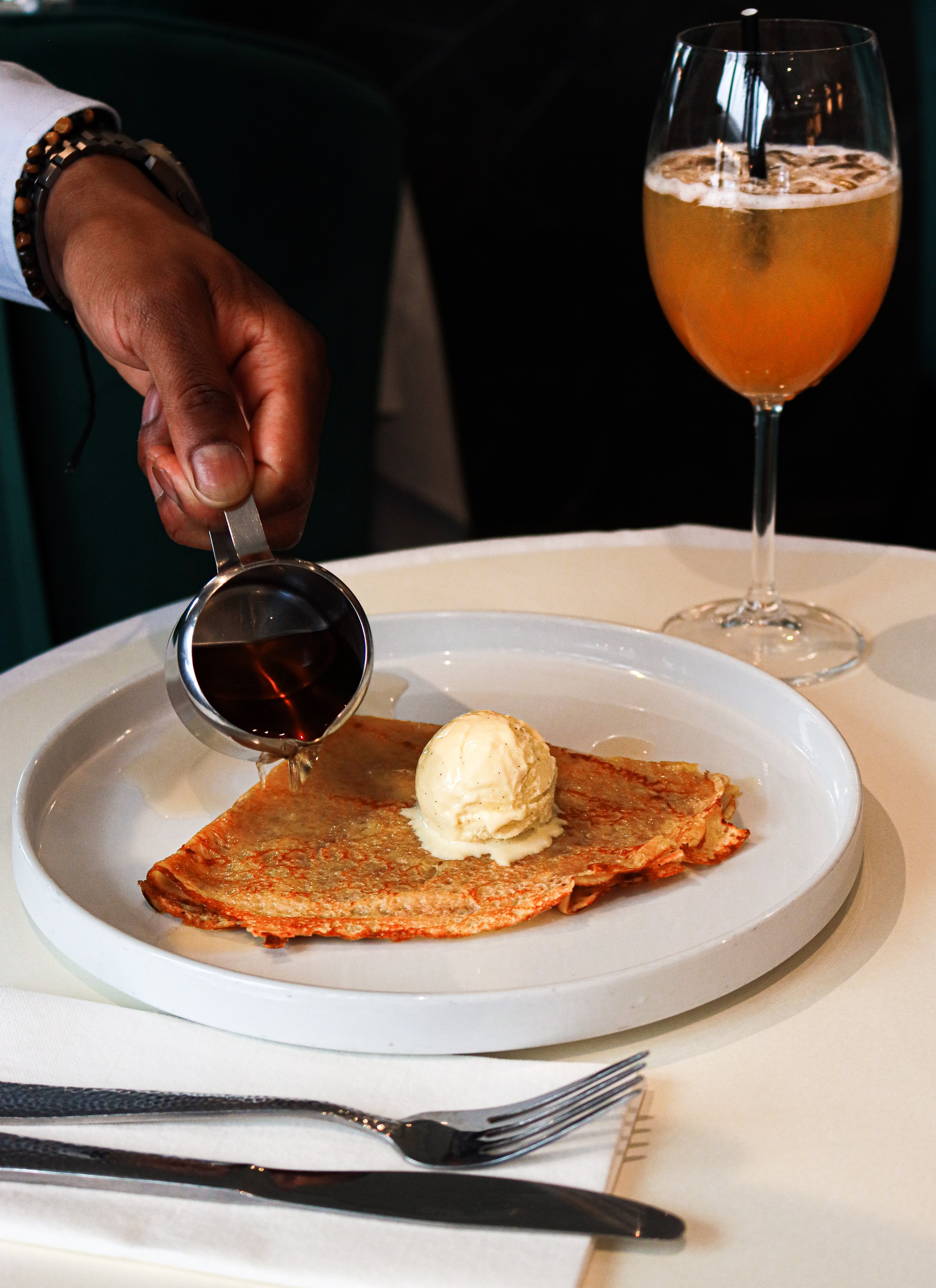 Assiette avec une crêpe fine, une boule de glace à la vanille, du sirop versé dessus, une garniture en arrière-plan, un verre de cocktail jaune avec un straw, une main tenant une petite louche avec du sirop.