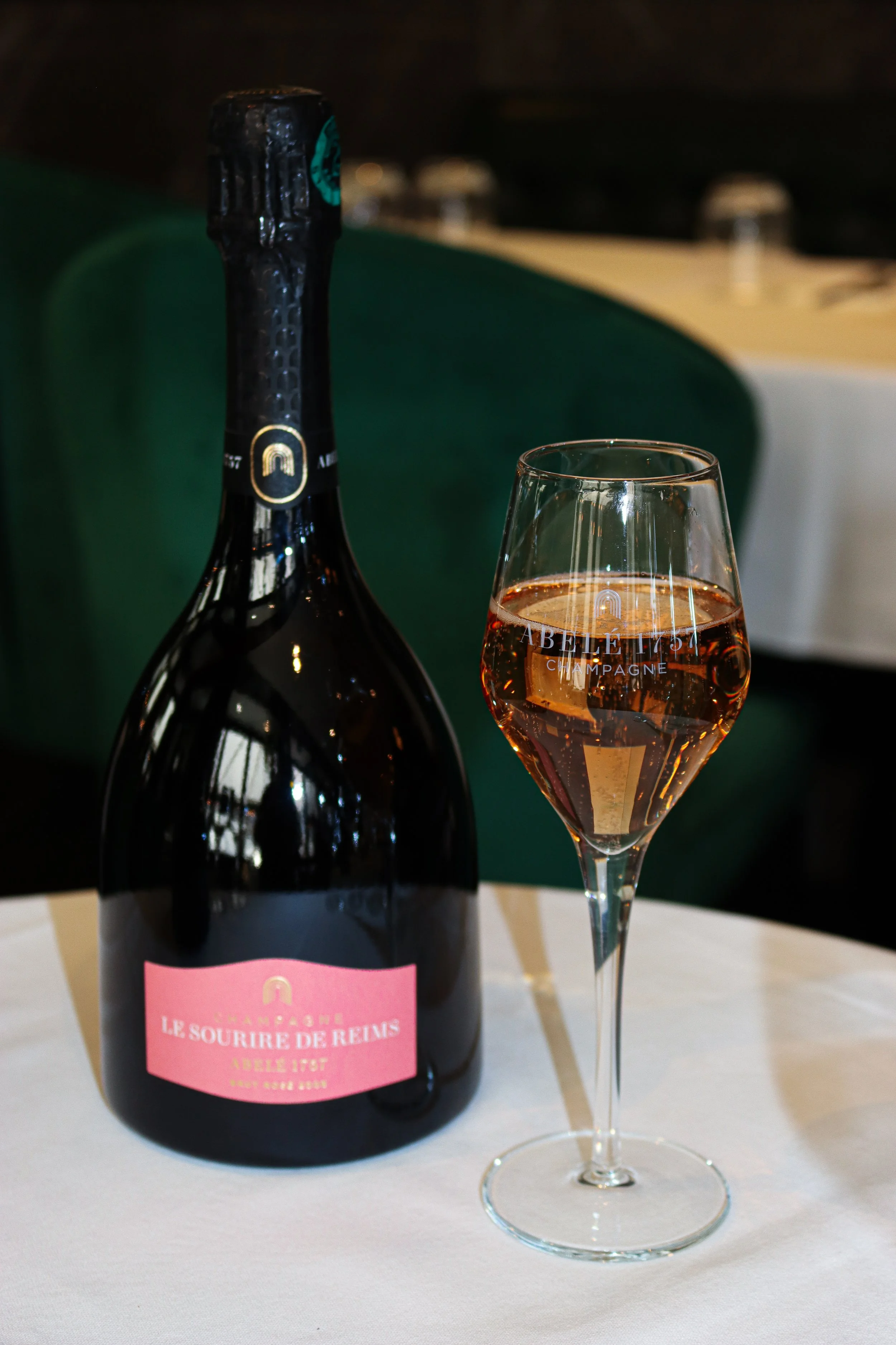 Une bouteille de champagne avec une étiquette rose et un verre de champagne rosé sur une table. L'arrière-plan est flou, avec une chaise verte en arrière-plan.