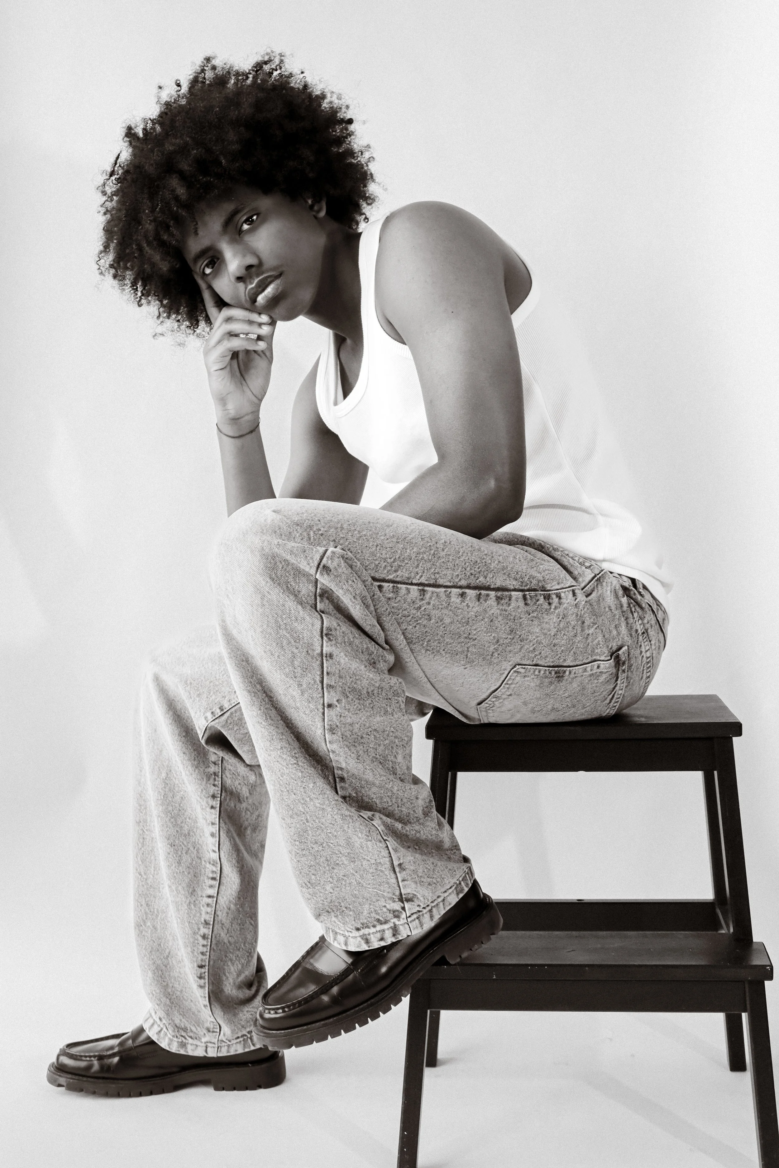 Une personne assise sur une chaise en bois, portant un t-shirt blanc, des jeans et des chaussures noires, regarde devant avec une expression sérieuse, avec la tête reposant sur sa main droite, dans une photo en noir et blanc.