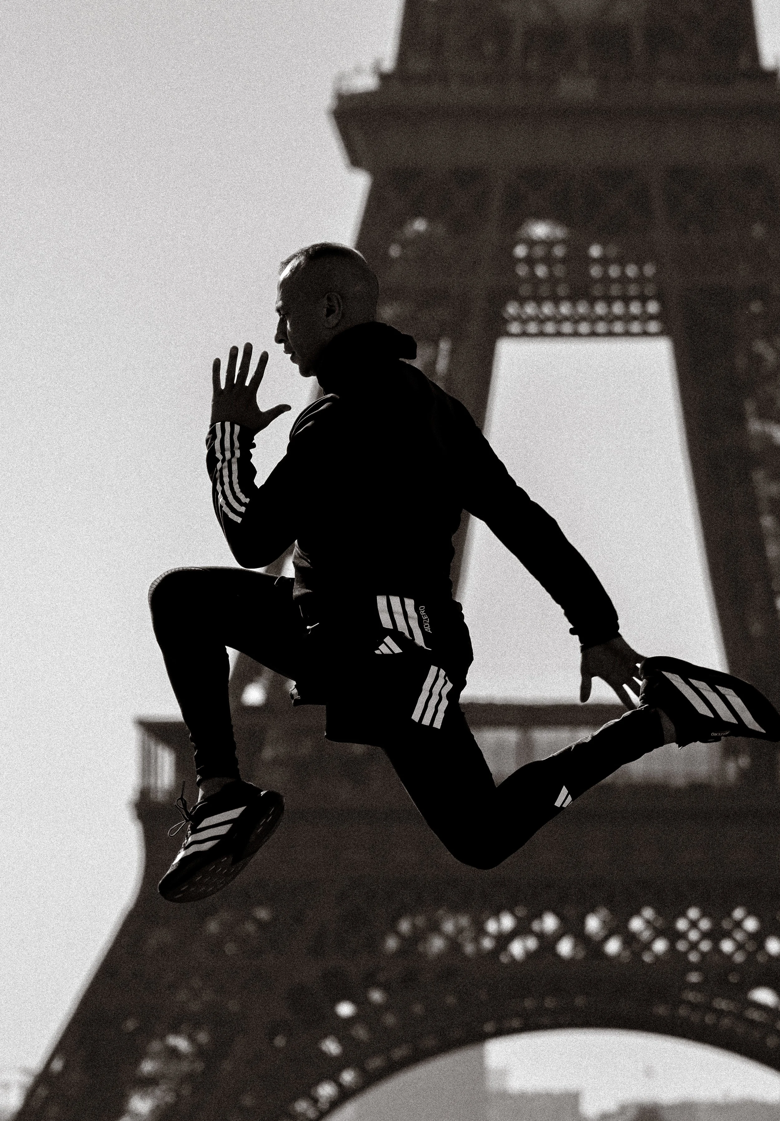Une personne en pleine course, sautant dans les airs devant la Tour Eiffel, photographiée en noir et blanc.