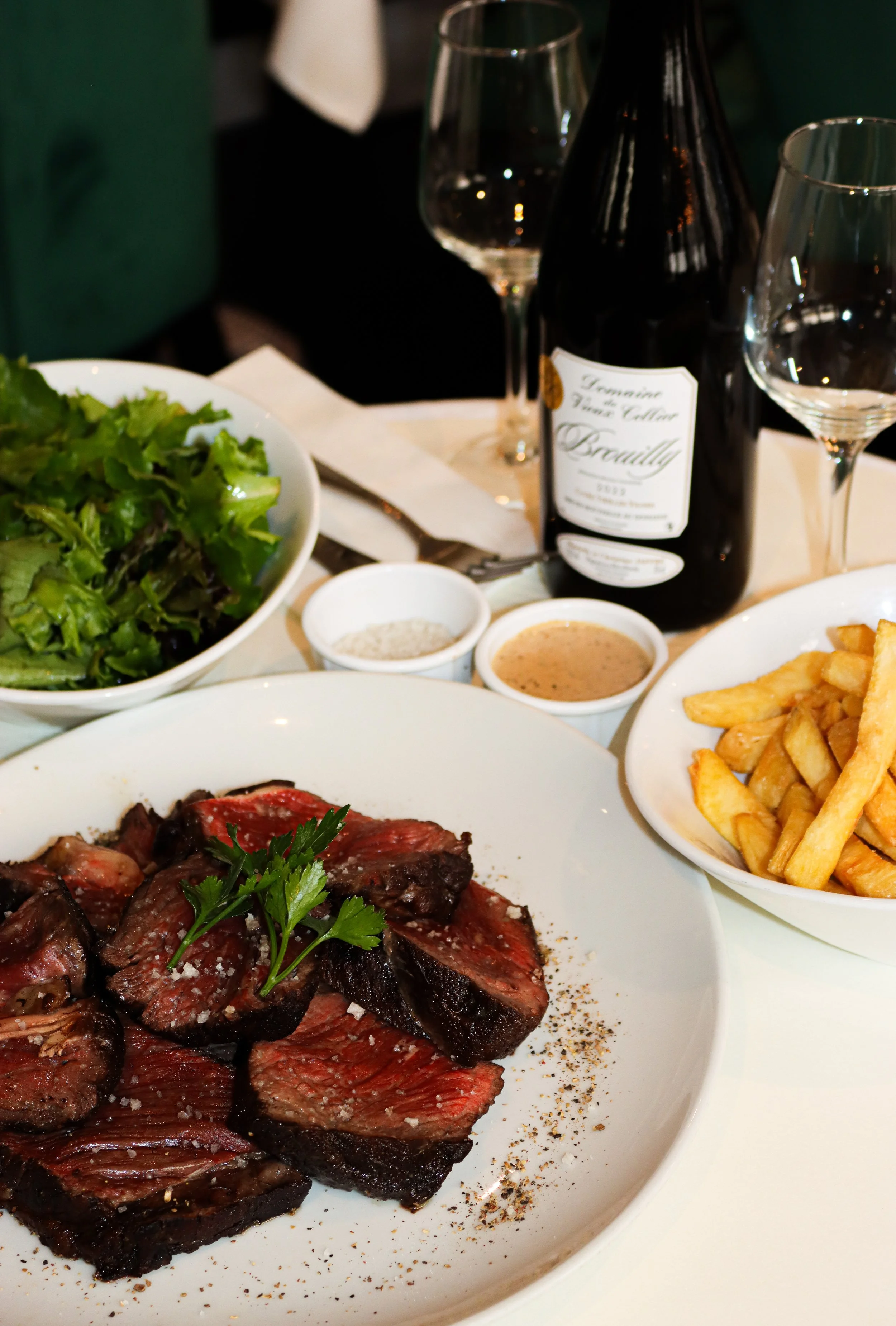 Un plat de viande saignante avec quelques morceaux de rosbif, accompagnée de frites, une salade verte, des sauces et une bouteille de vin sur la table.